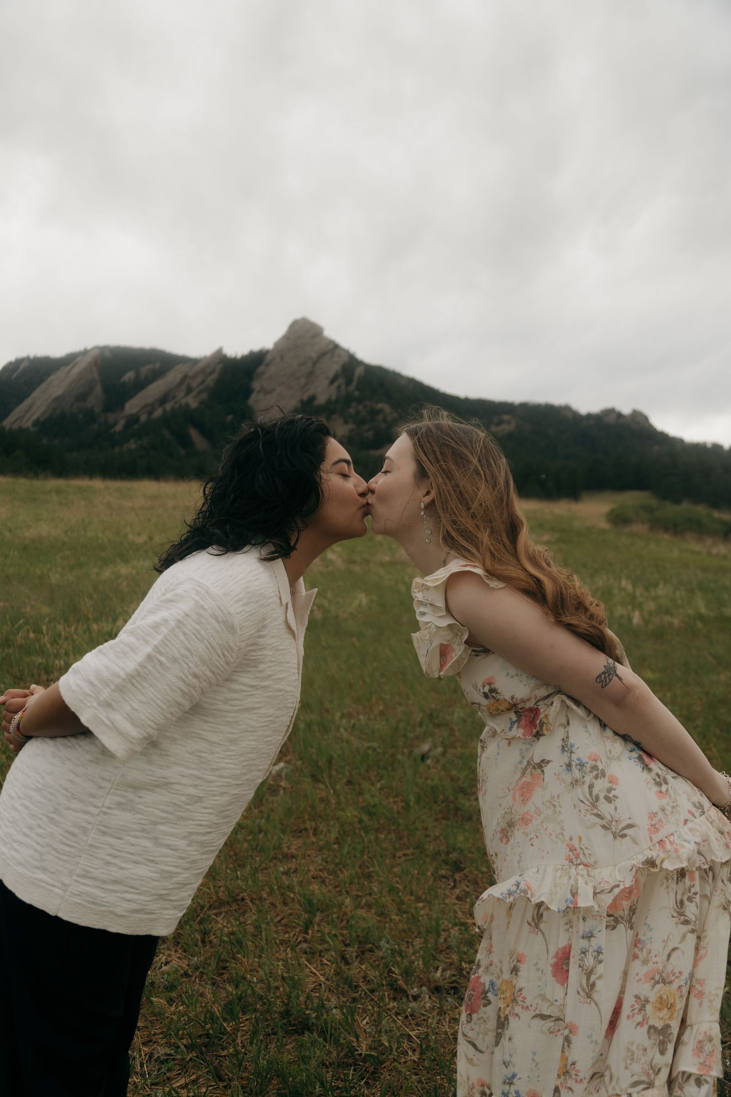 Two women share a romantic kiss in a grassy field with dramatic rocky mountain flatirons behind them.