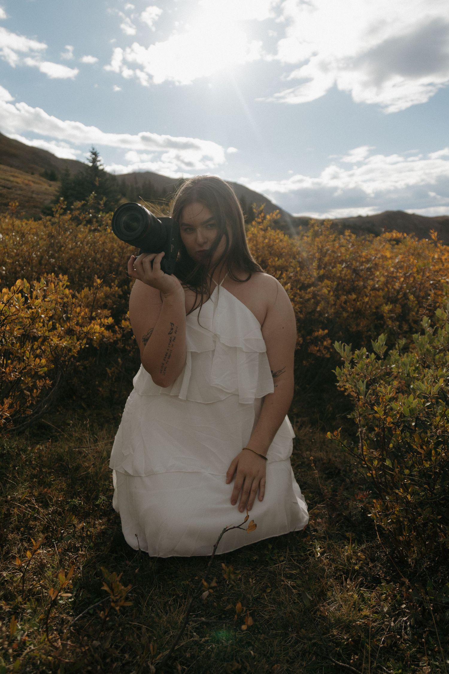 Woman in white dress kneeling in golden field, holding camera up, with sunlit mountains in background.