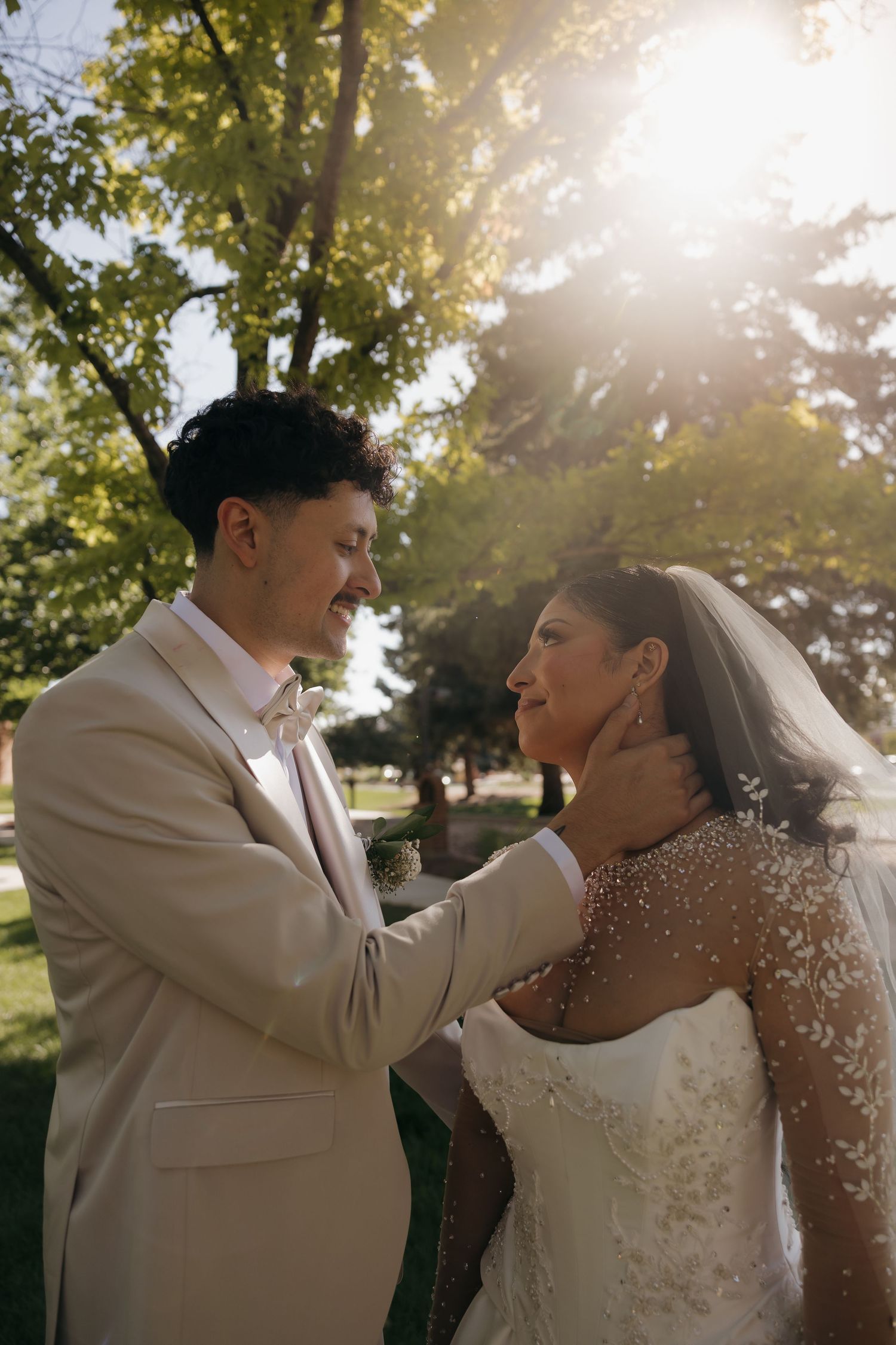 Groom in beige suit gently holds brides face in sunlit outdoor wedding portrait.