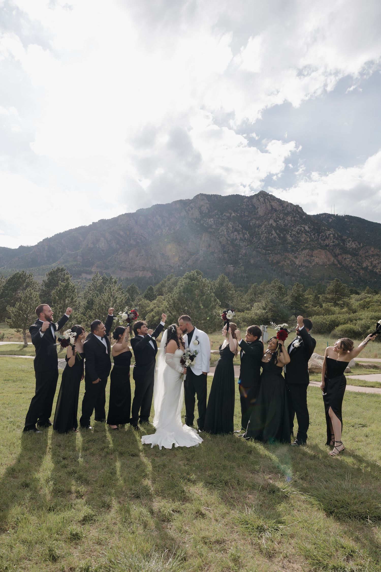 Wedding party in black attire celebrating with bride and groom kissing against mountain backdrop.