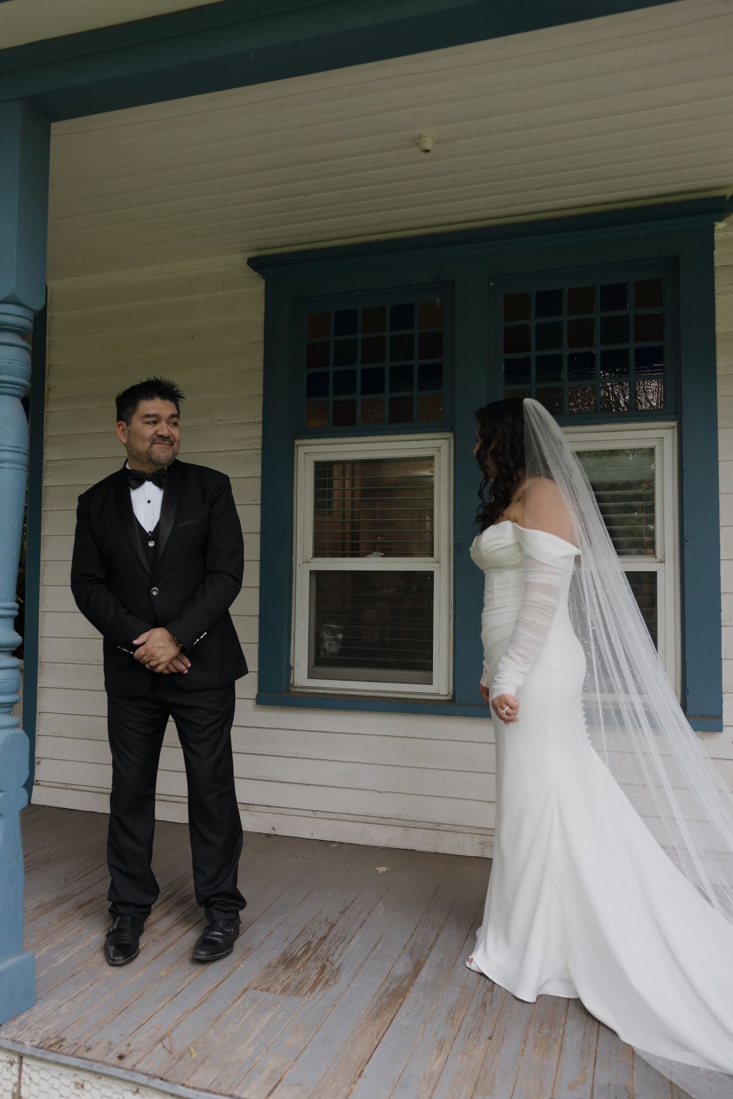 A groom in a black tuxedo smiles at his bride in a white off-shoulder gown and veil on a wooden porch.