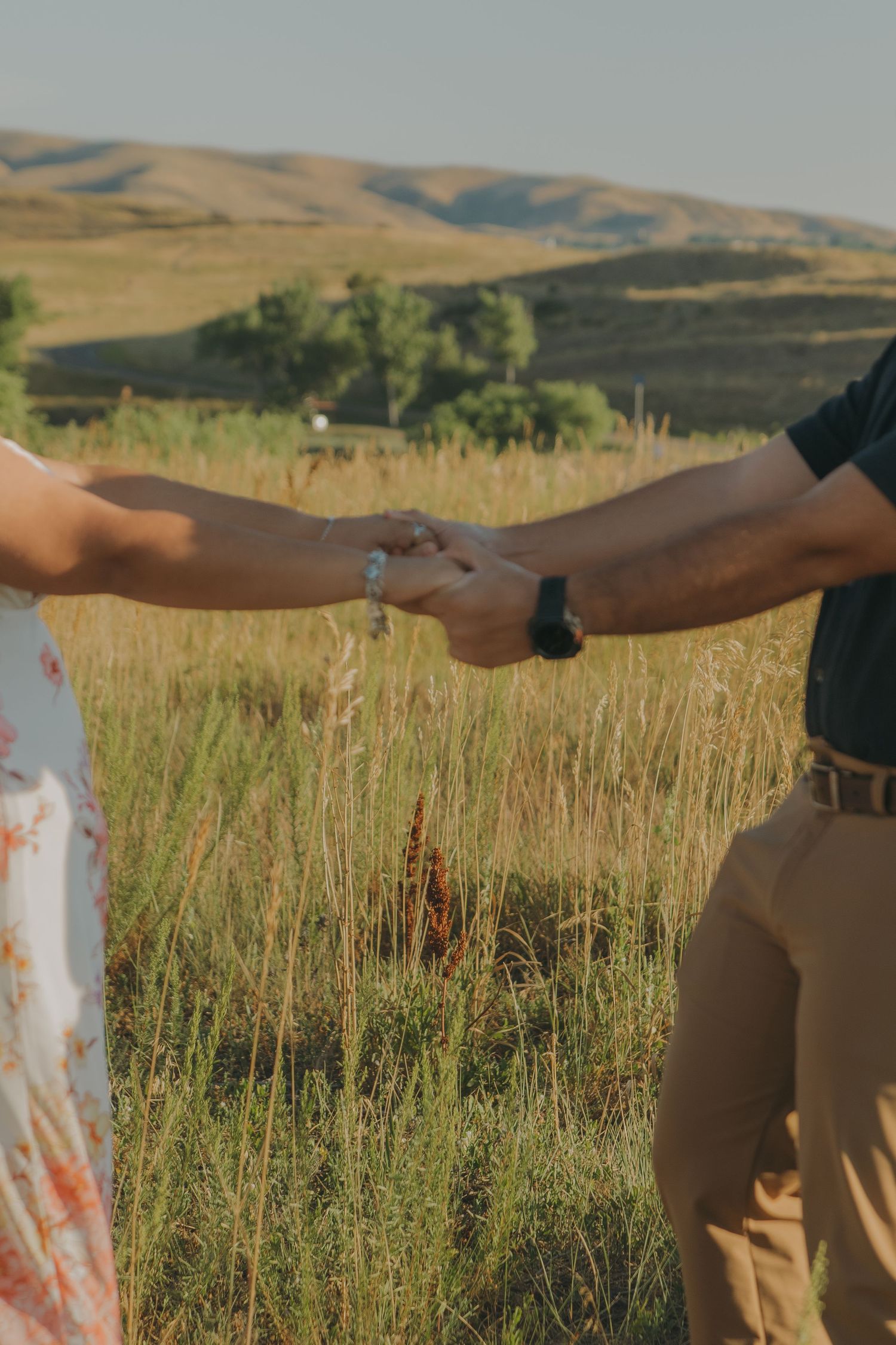 Two people hold hands in a scenic field with golden hills stretching into the distance.