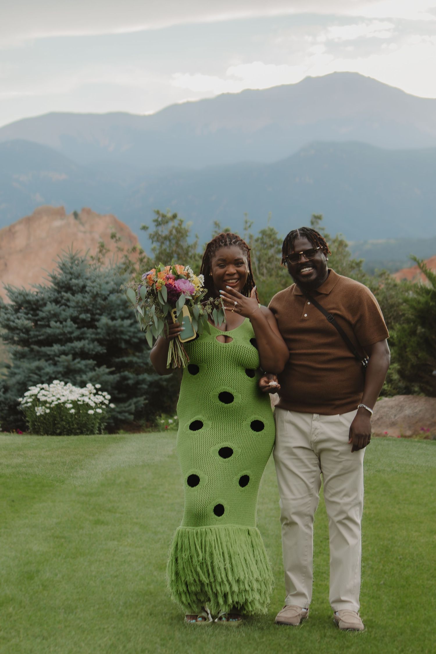 A couple poses outdoors in front of mountains, wearing a green polka dot dress and brown polo shirt.