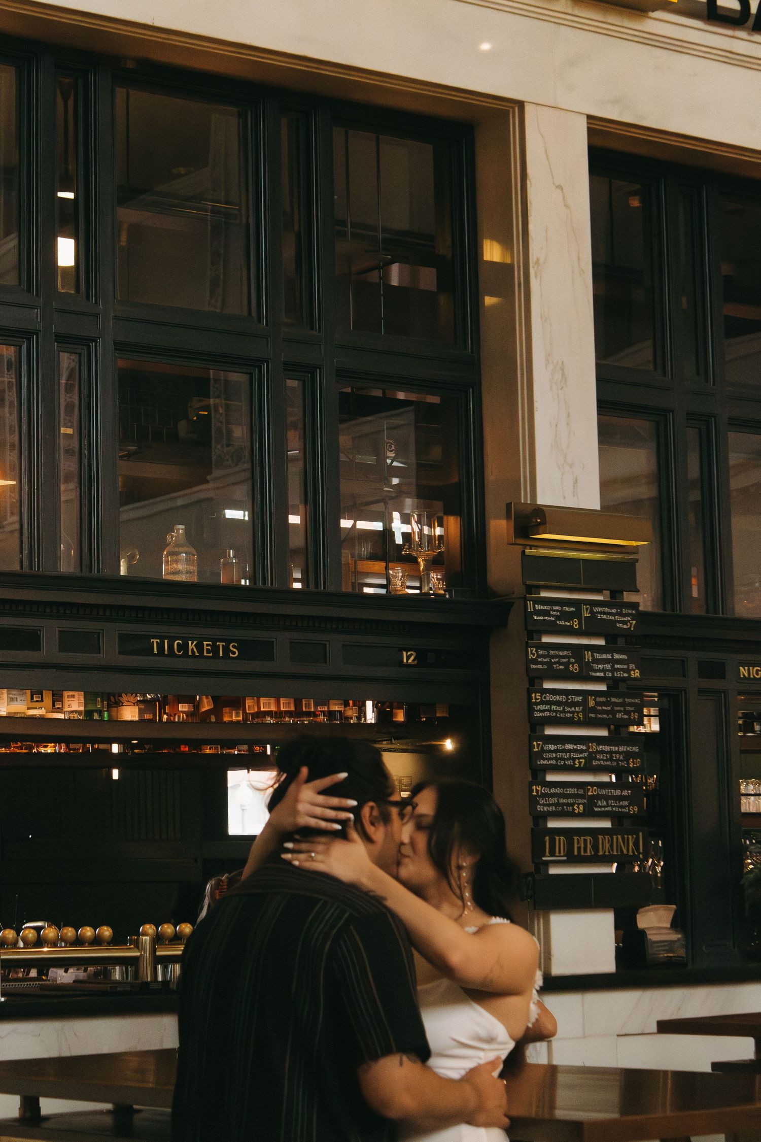 Romantic couple embrace in a dimly lit restaurant with large windows and dark wood paneling.