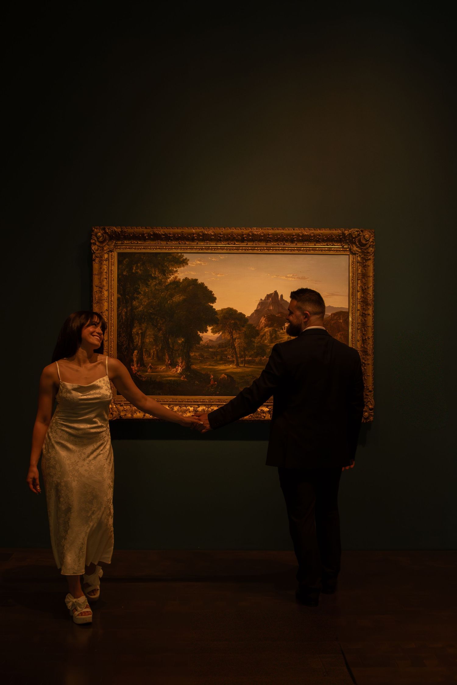 Dimly lit art gallery scene showing people viewing large landscape paintings on dark walls.