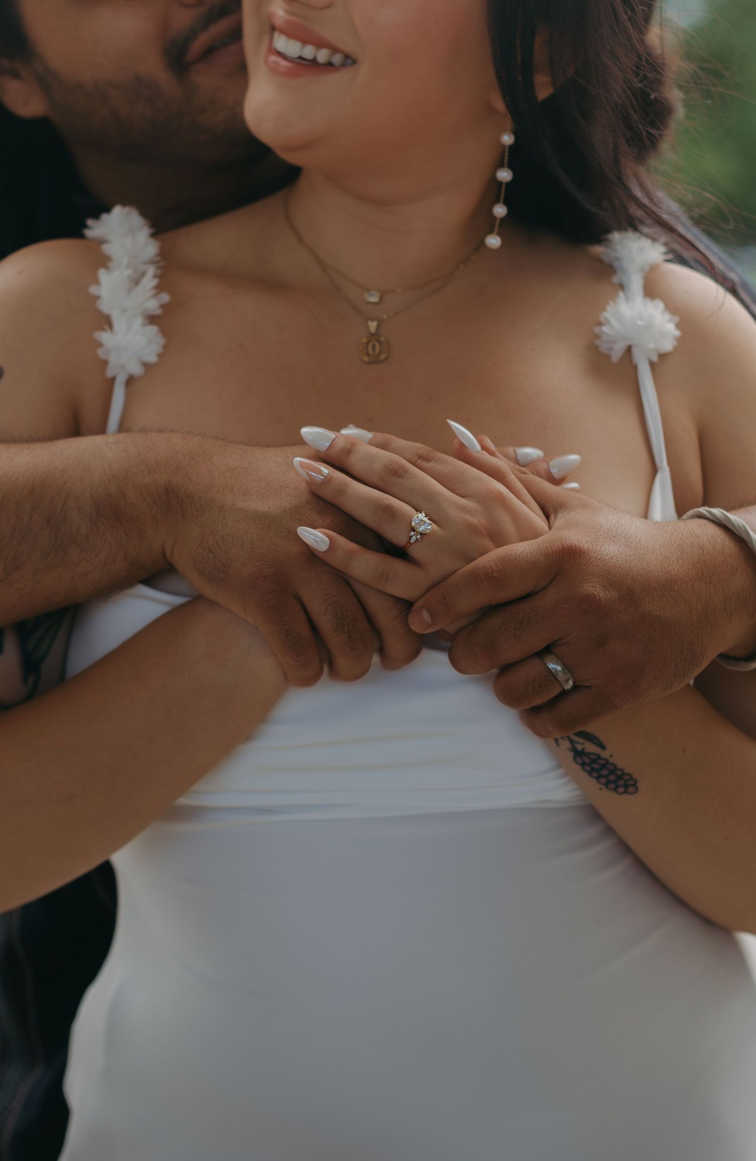 Close up of hands with wedding rings and white dress details showing pearl necklace and floral straps.