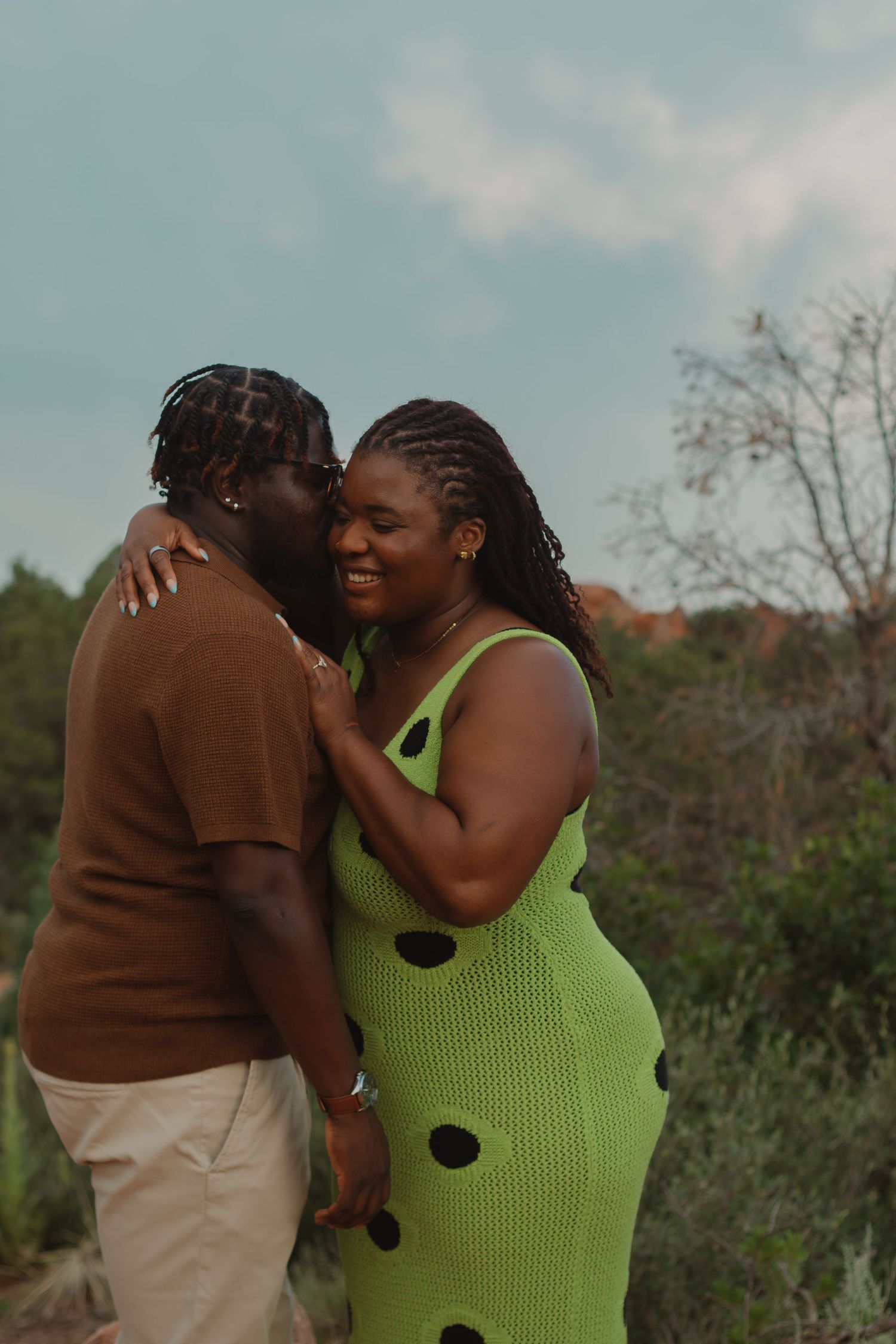 A loving couple embraces outdoors against a scenic mountain backdrop on a sunny afternoon.