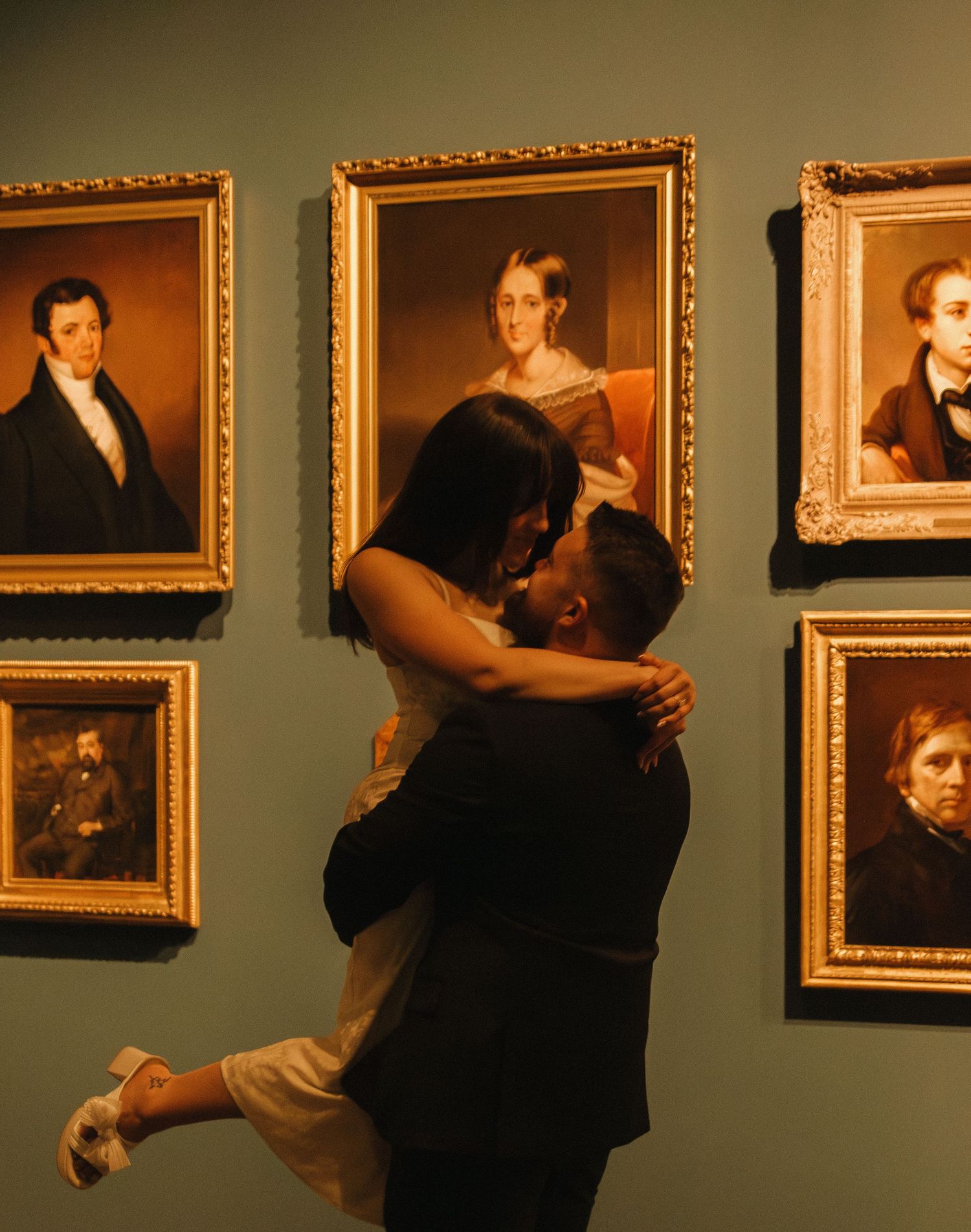 A tender embrace shared in an art gallery surrounded by historic portrait paintings in gilded frames.
