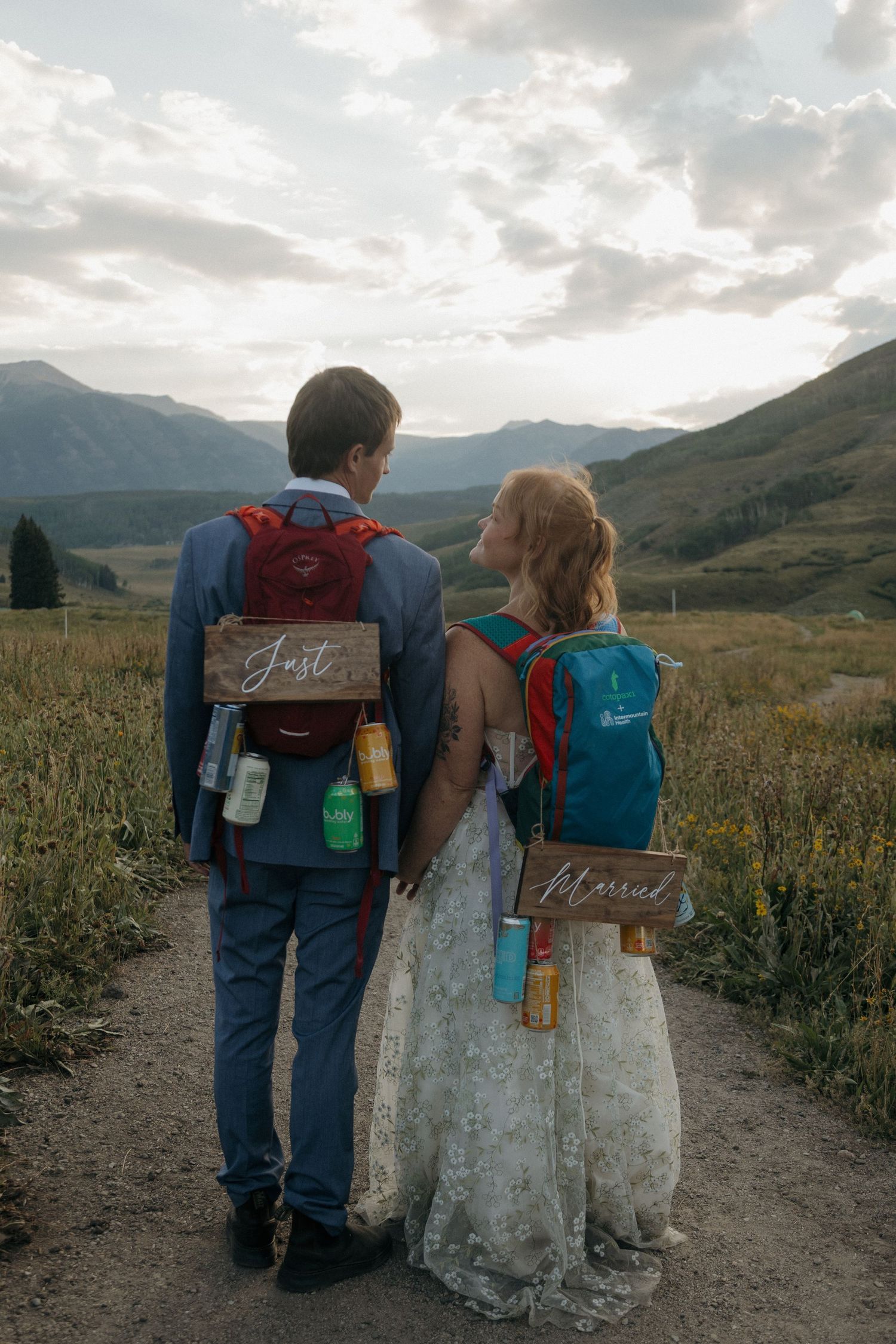 Bride and groom with hiking backpacks labeled Just Married walking toward mountain landscape at sunset.