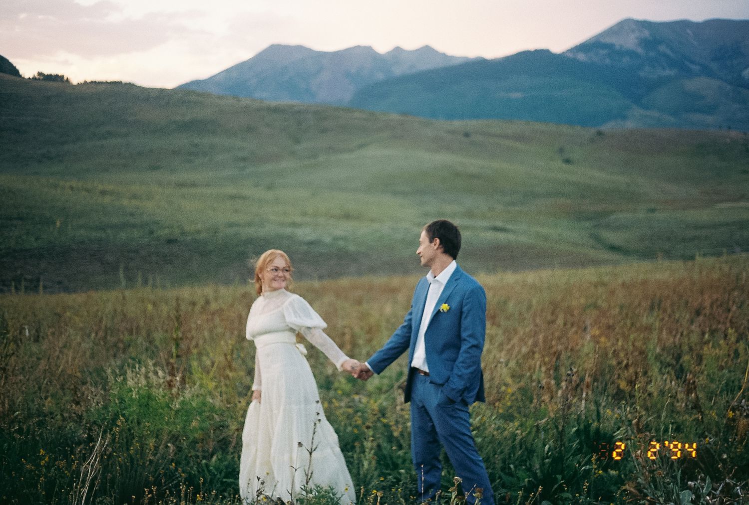 Bride and groom holding hands in a lush green mountain meadow at sunset during their wedding.