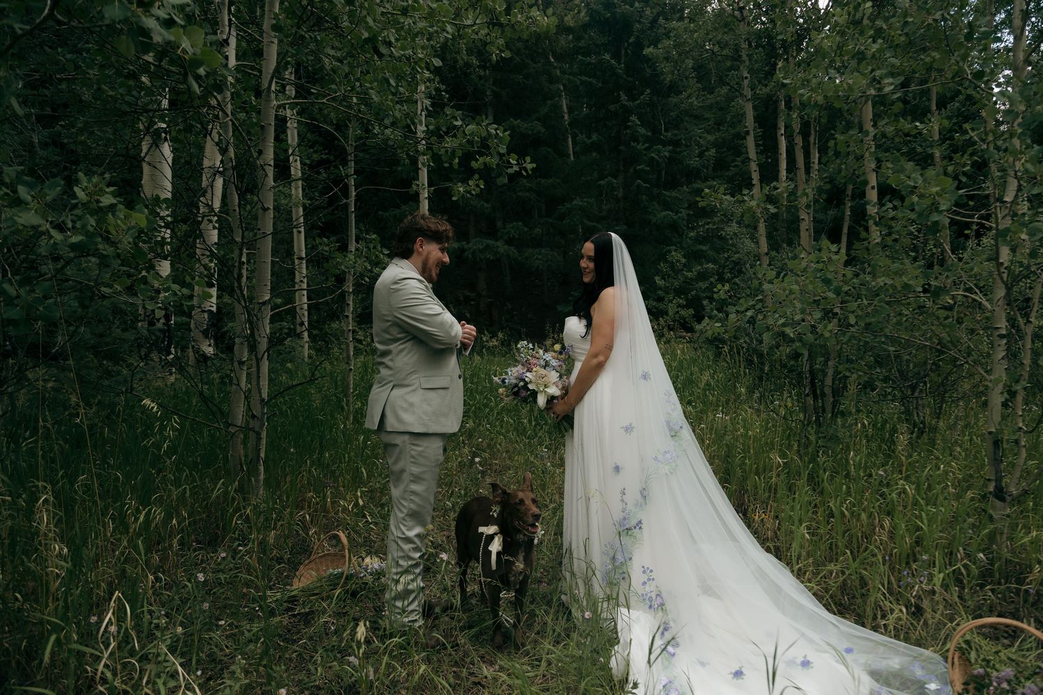 Bride and groom exchange vows in a lush forest setting, with a black dog beside them, bride wearing a flowing veil.