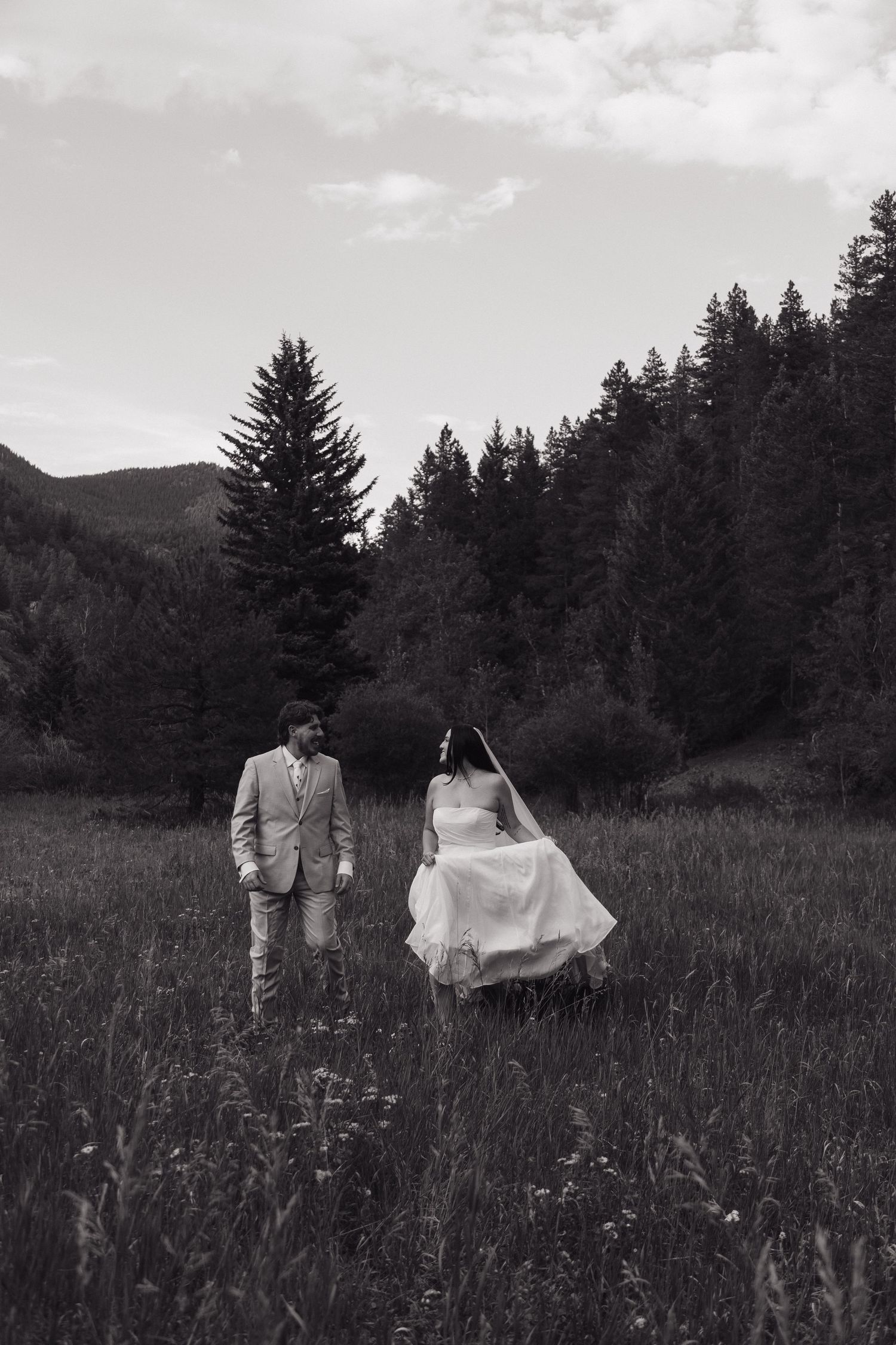 Black and white photo of bride and groom walking through wildflower meadow with mountain forest behind.