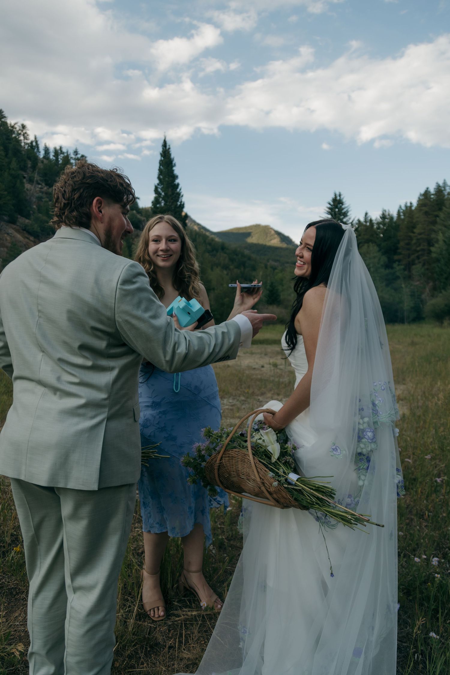 Bride and groom celebrate with an officiant in a mountain meadow, sharing a toast during their outdoor elopement ceremony.
