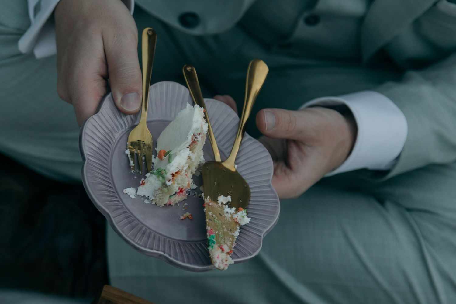Close-up of groom holding two plates of wedding cake slices with gold forks.
