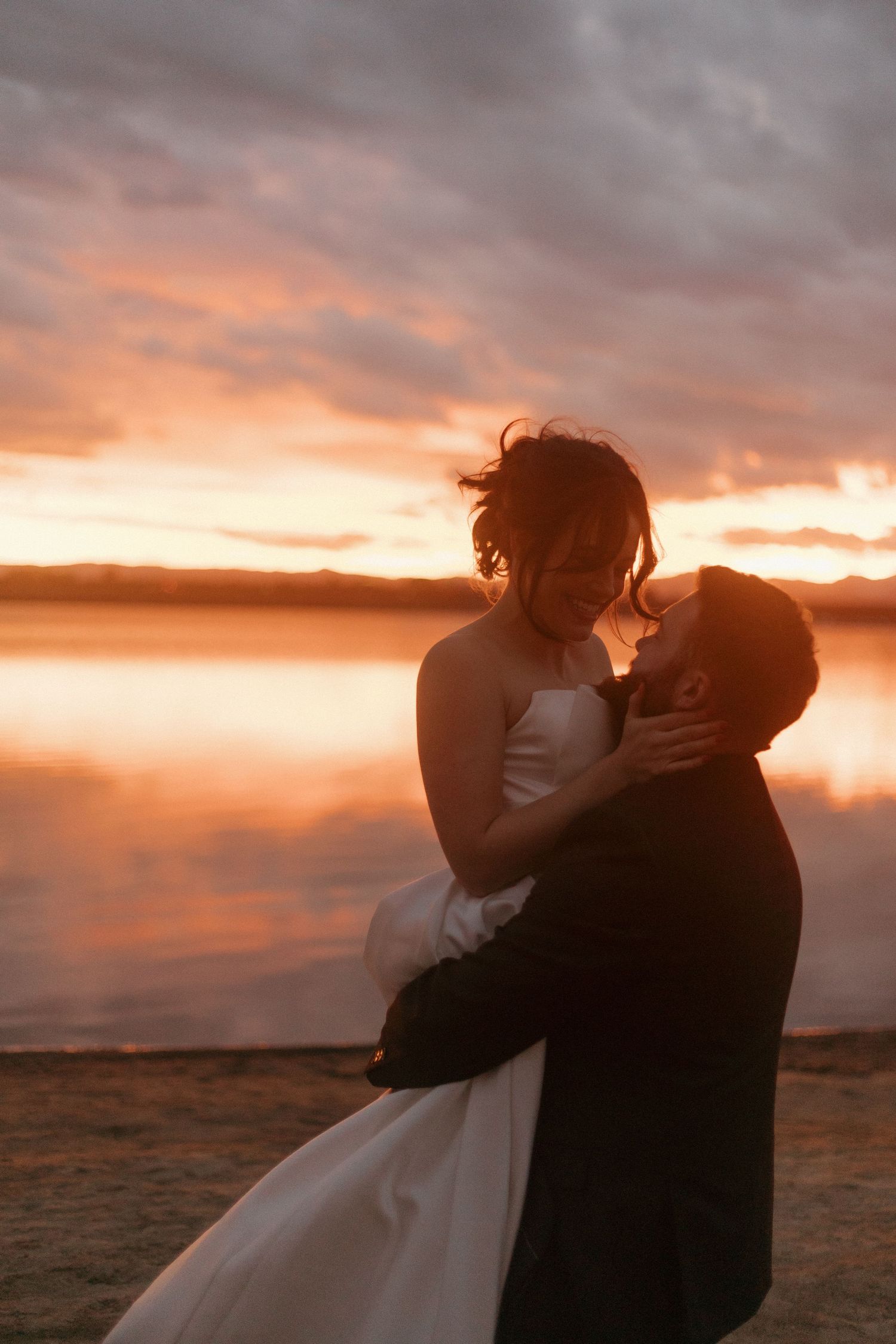 Silhouetted couple in wedding attire sharing a romantic kiss against a dramatic sunset over water.