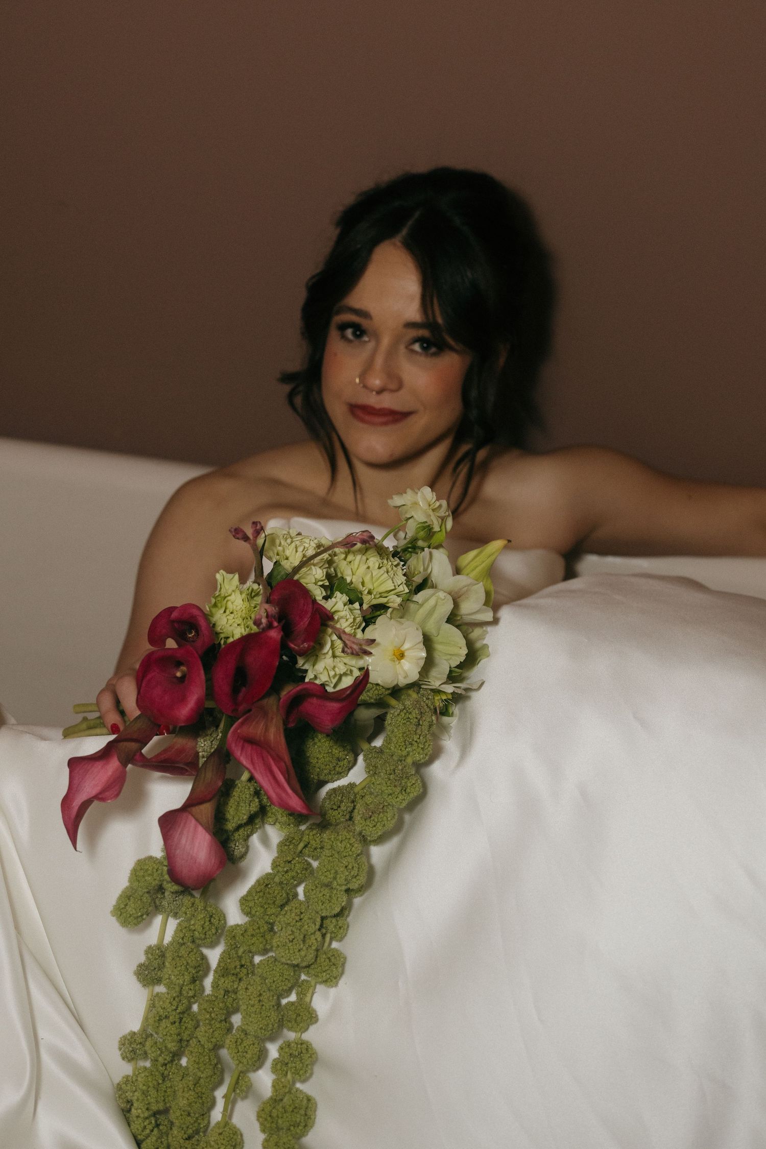 Artistic bridal portrait with red roses and green floral arrangements against white bedding.