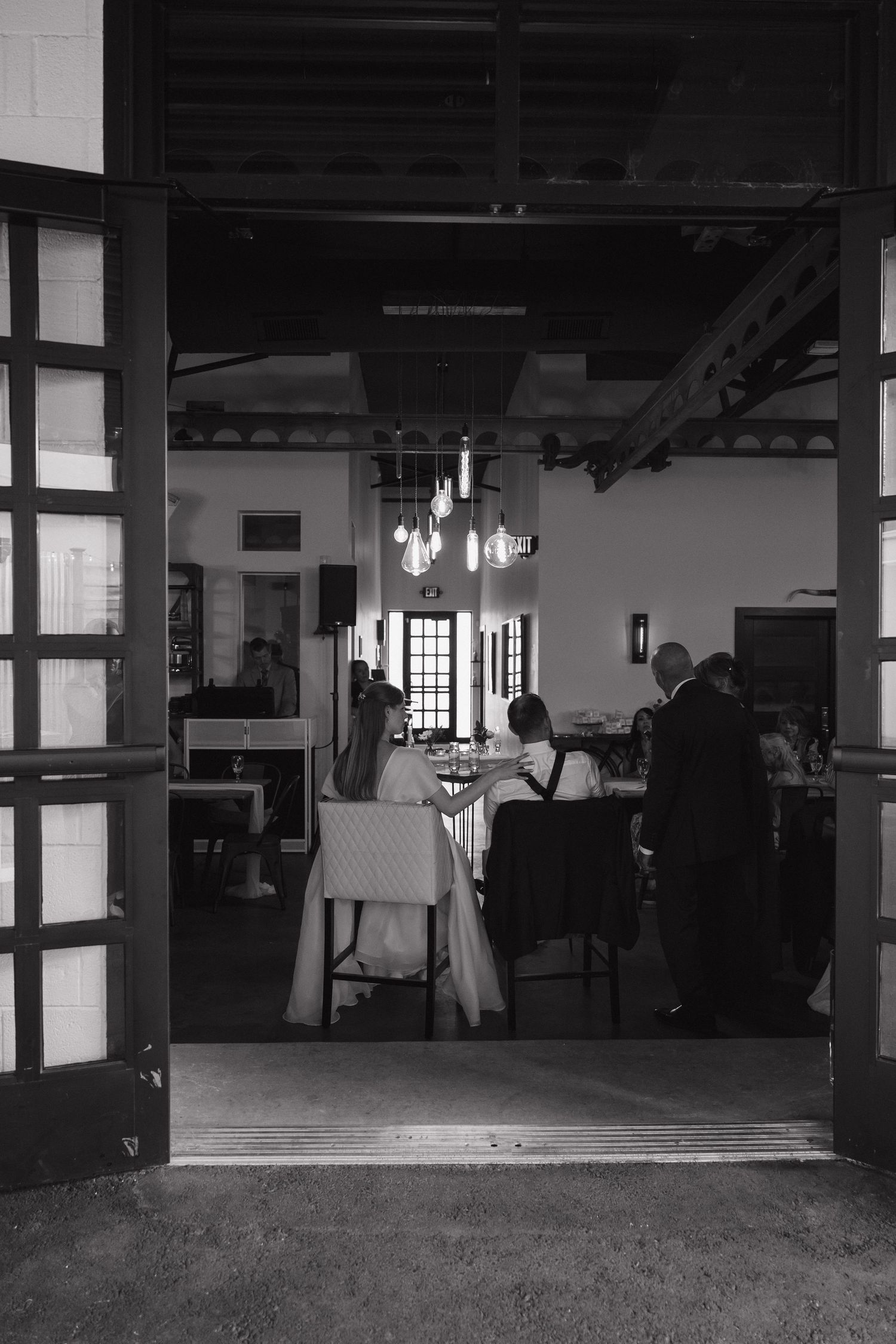 Bride and groom share a quiet moment at their reception table, viewed through elegant open wooden doors.