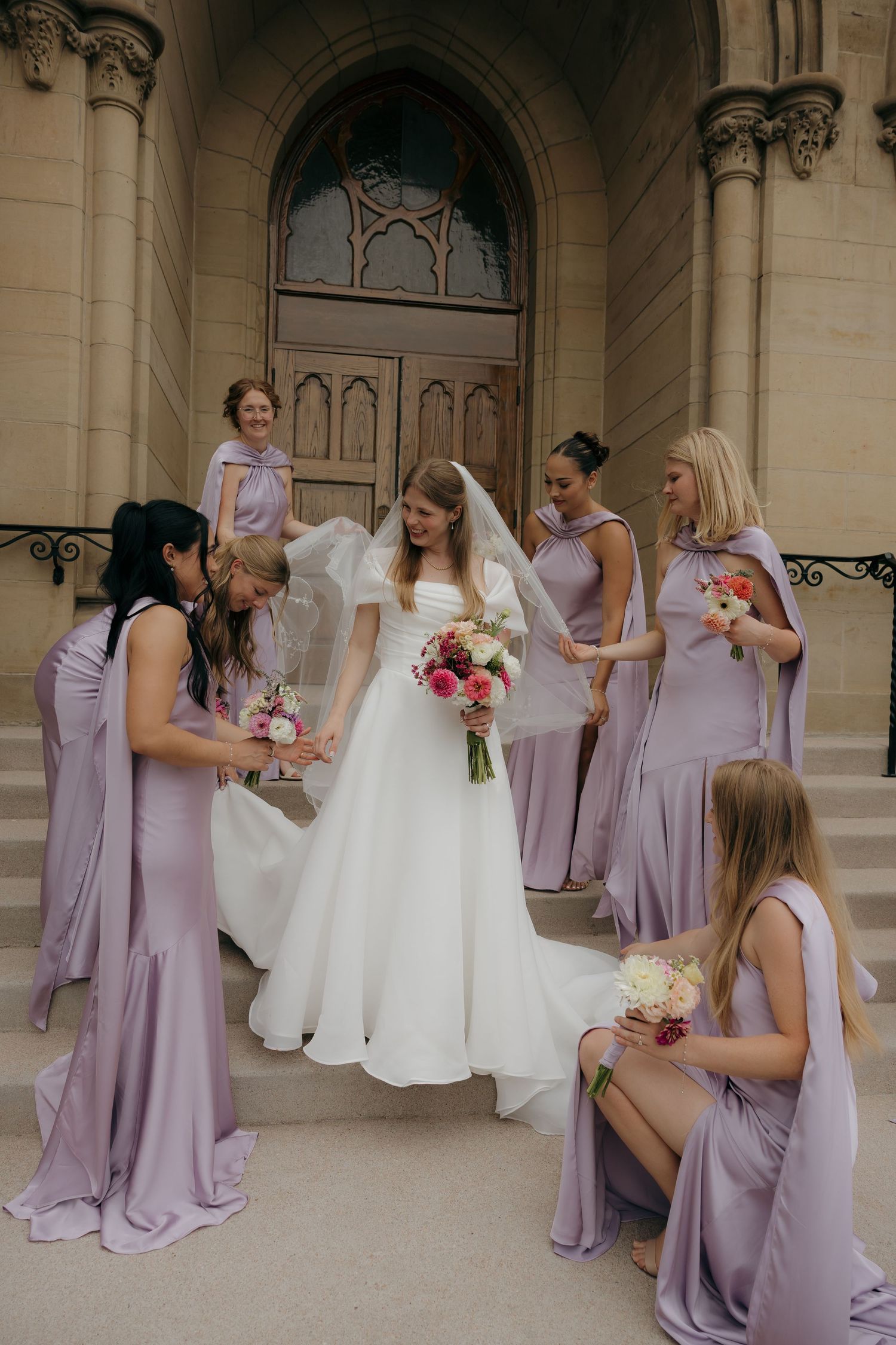 Bride in white gown surrounded by bridesmaids in lavender dresses on church steps.