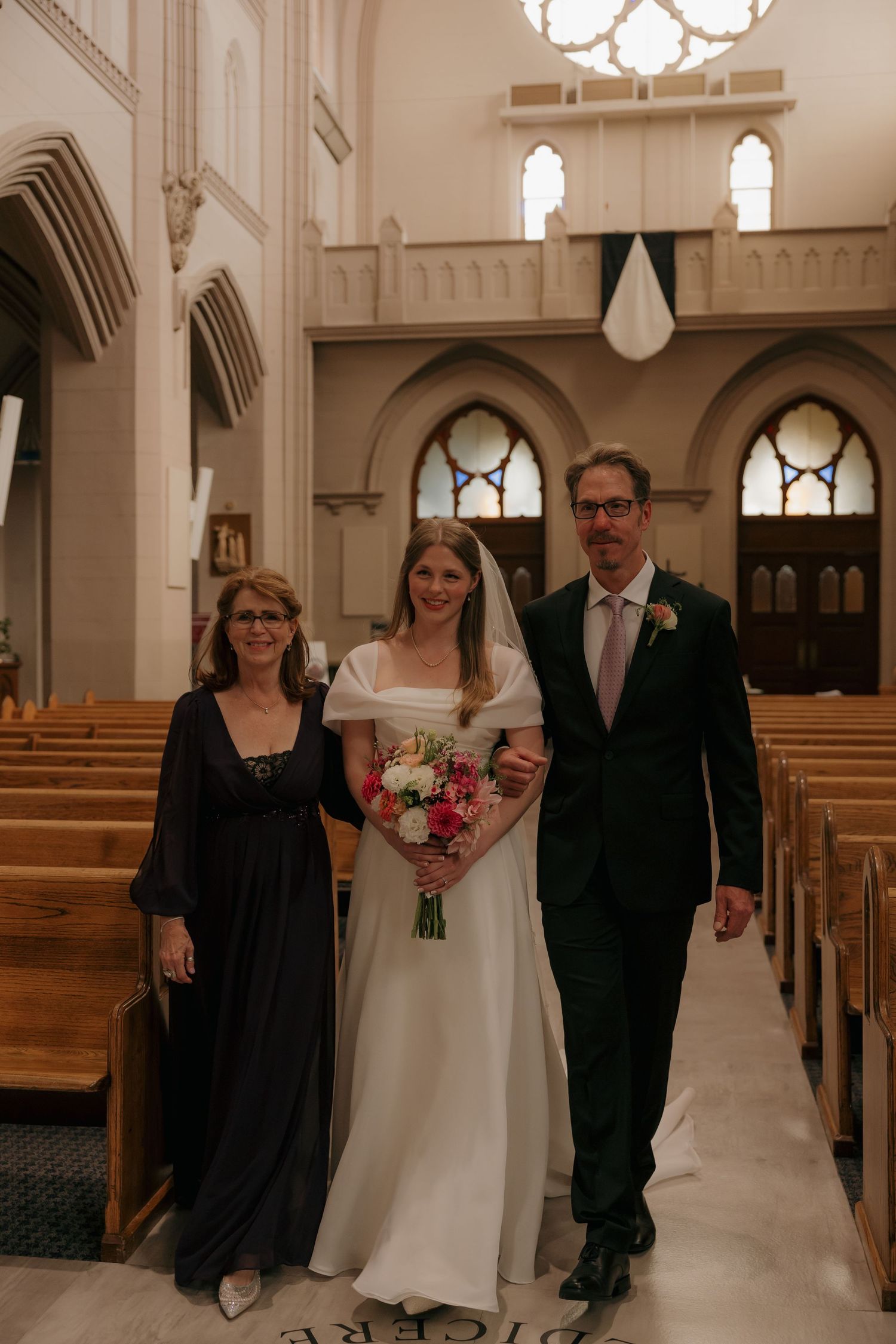 Bride in white off-shoulder gown holding pink bouquet walks down church aisle with parents.