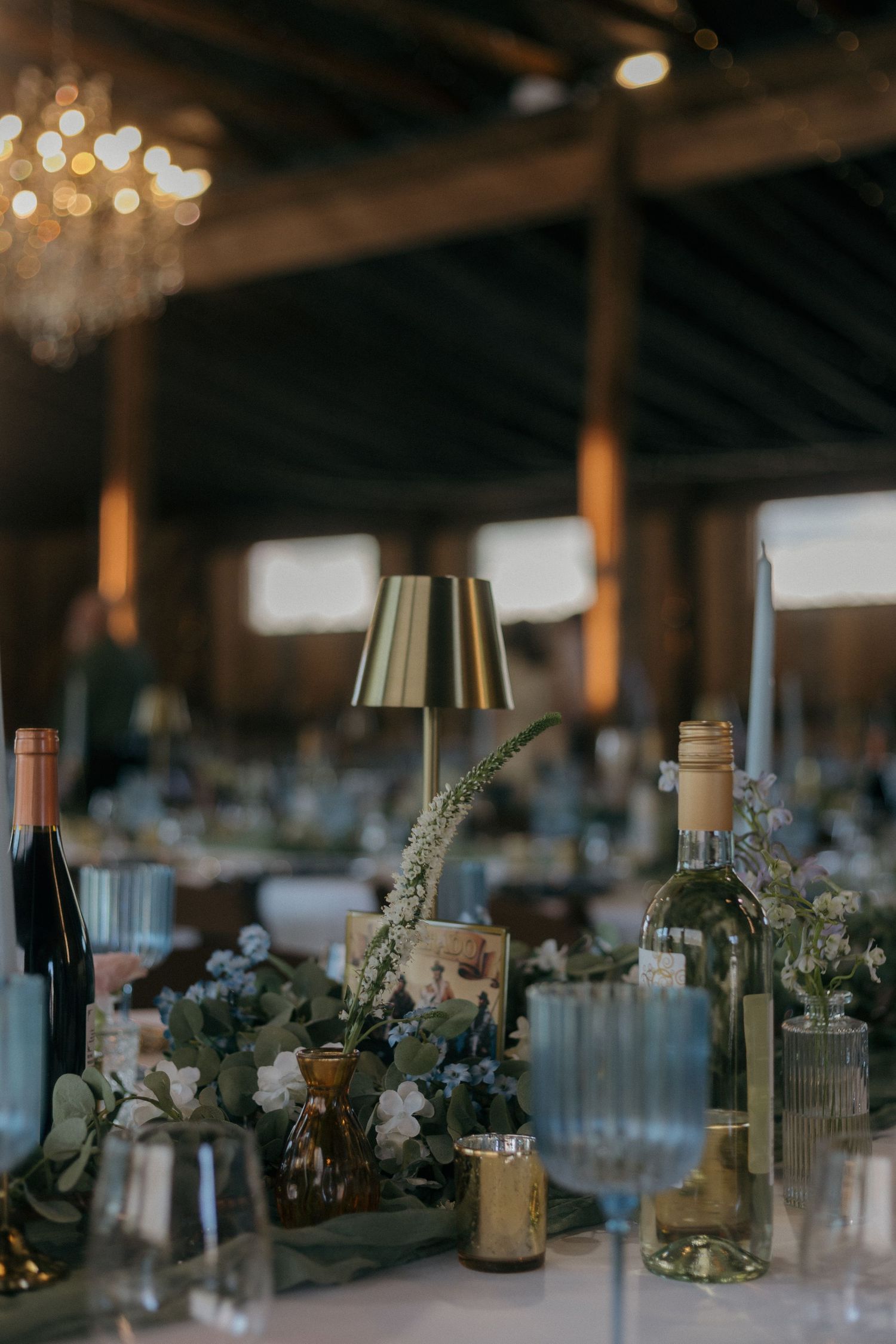Elegant wedding reception table with gold lamp, wine bottles, floral greenery, and crystal glassware in a rustic barn venue.