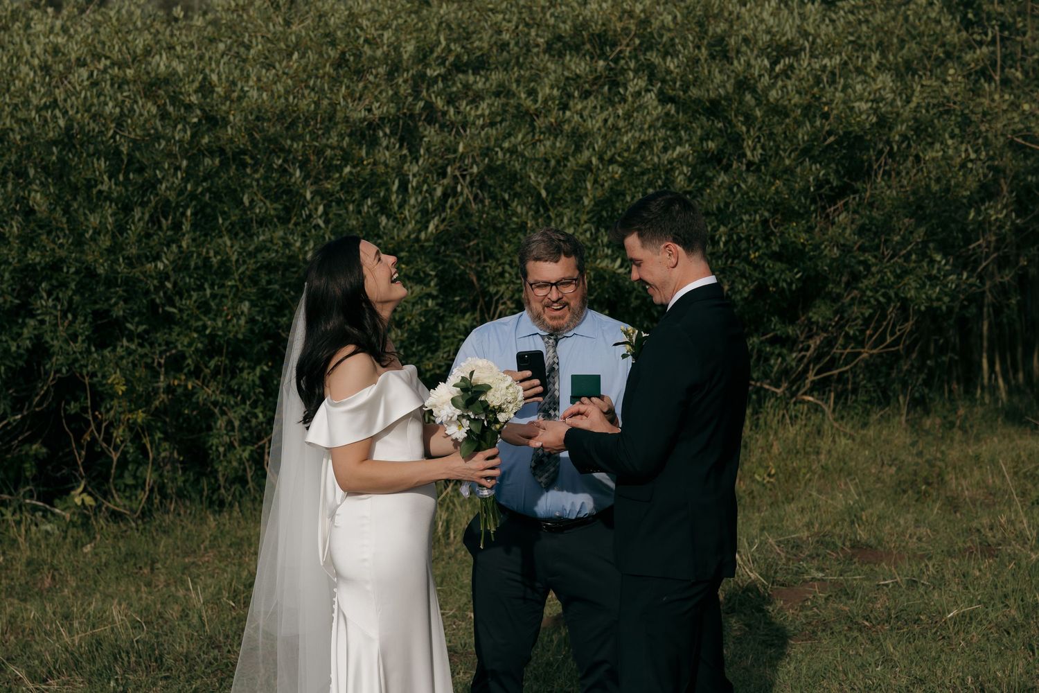 Bride and groom exchange vows outdoors with officiant holding phone, surrounded by lush green foliage.