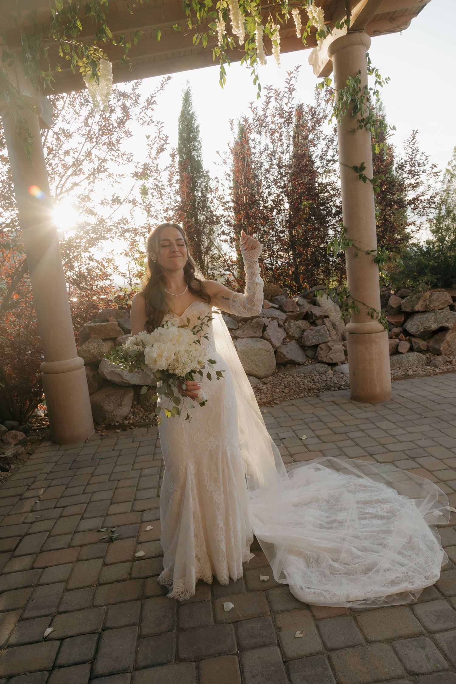 Radiant bride in lace gown with bouquet posing under pergola in golden sunlight.