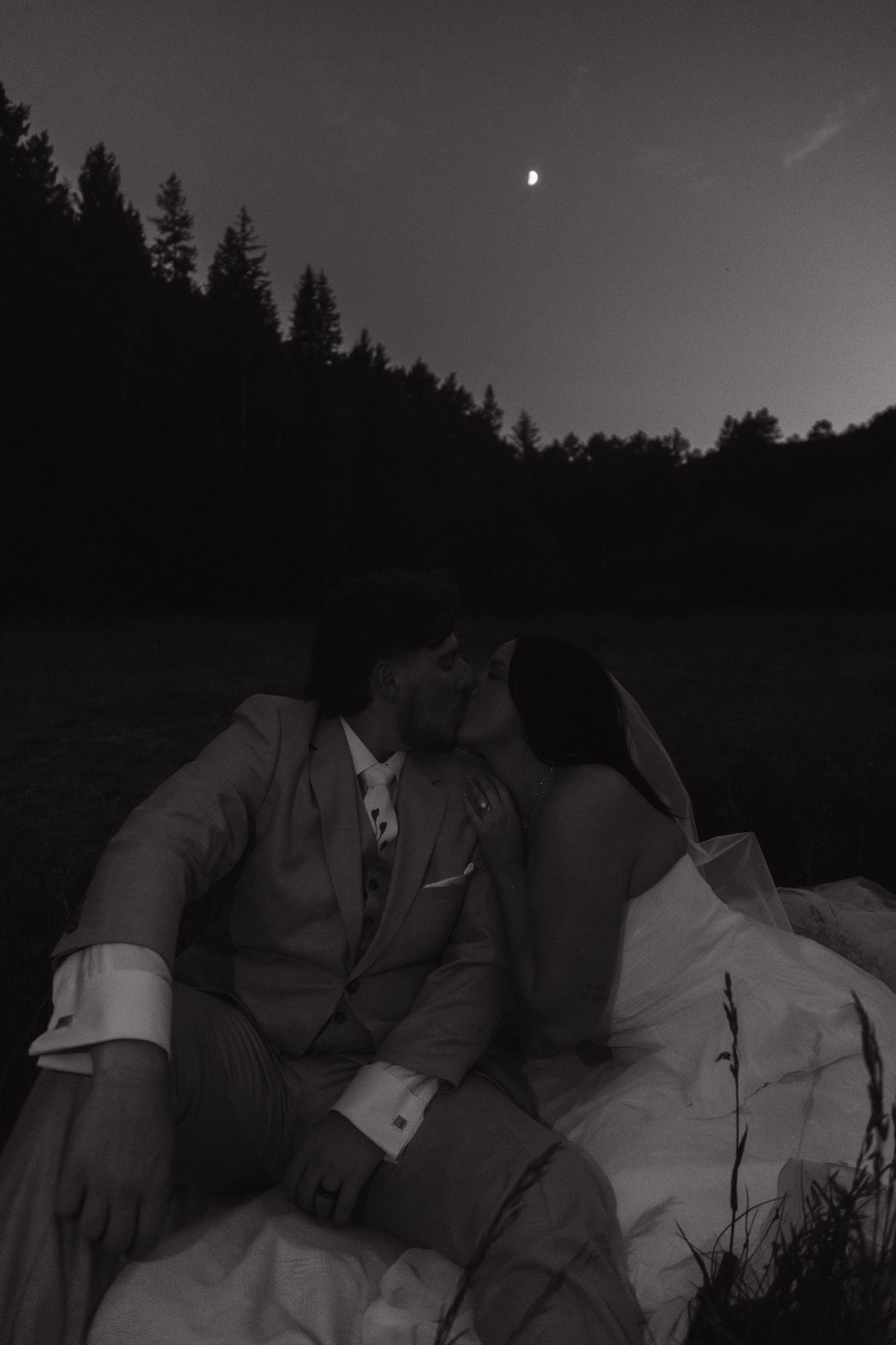 Black and white photo of bride and groom sharing a moonlit kiss outdoors, sitting together under a night sky with trees silhouetted.