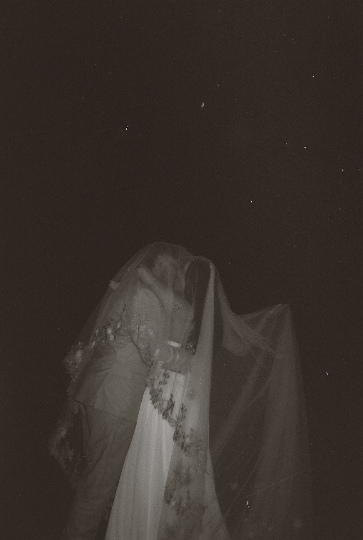 Dreamy dark night wedding portrait of couple kissing beneath a flowing floral lace veil.