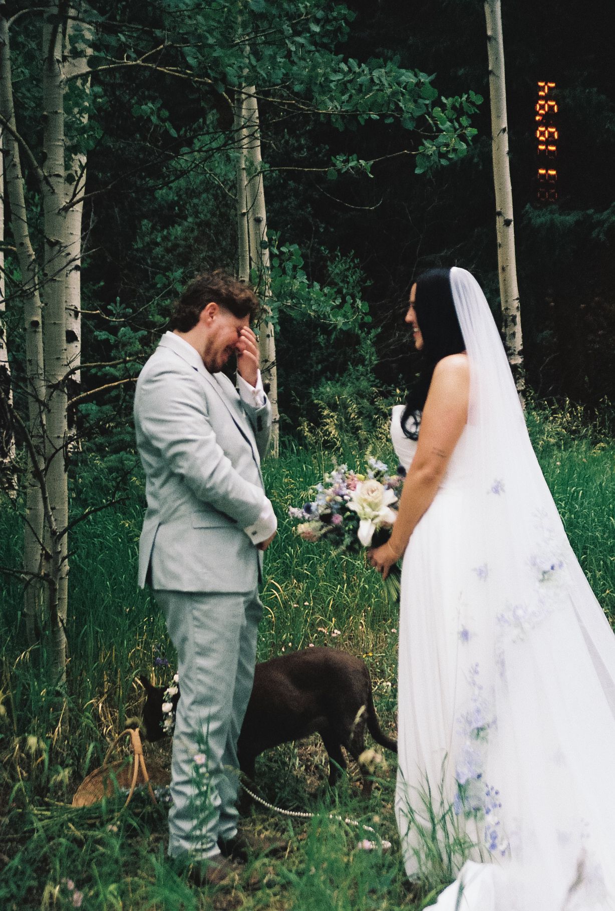 Emotional groom covering face in tears during first look with bride holding wildflower bouquet in aspen forest setting.