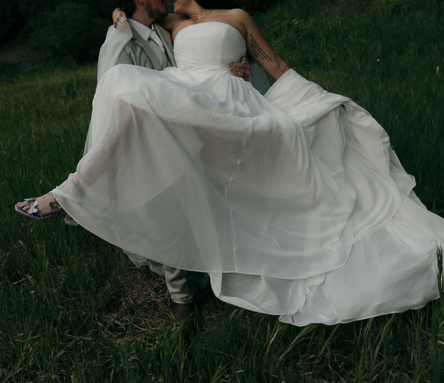 Bride and groom embrace in a green meadow, her flowing white strapless wedding gown billowing in the breeze.