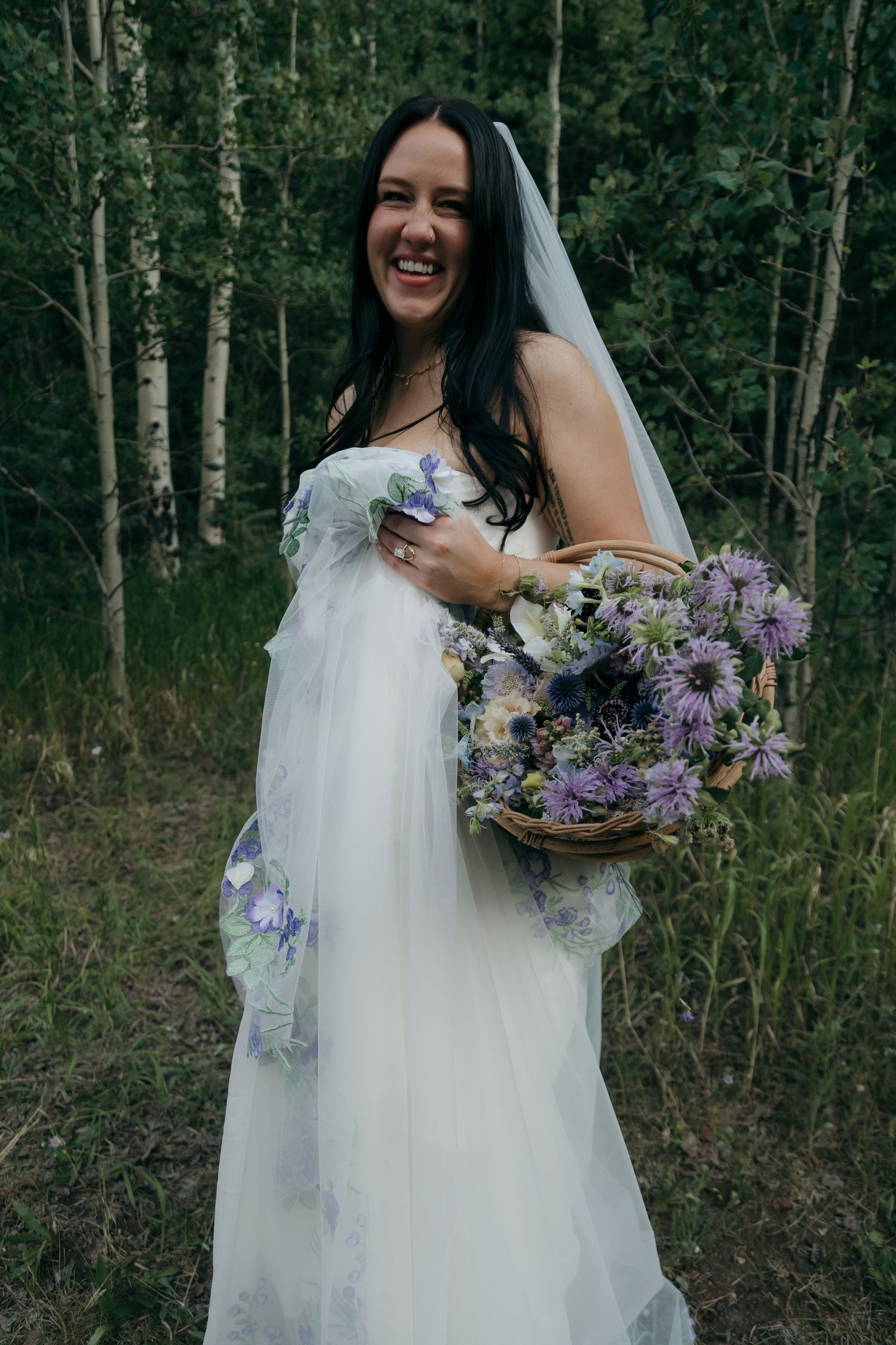 Smiling bride in floral wedding gown holds purple wildflower bouquet in birch forest.