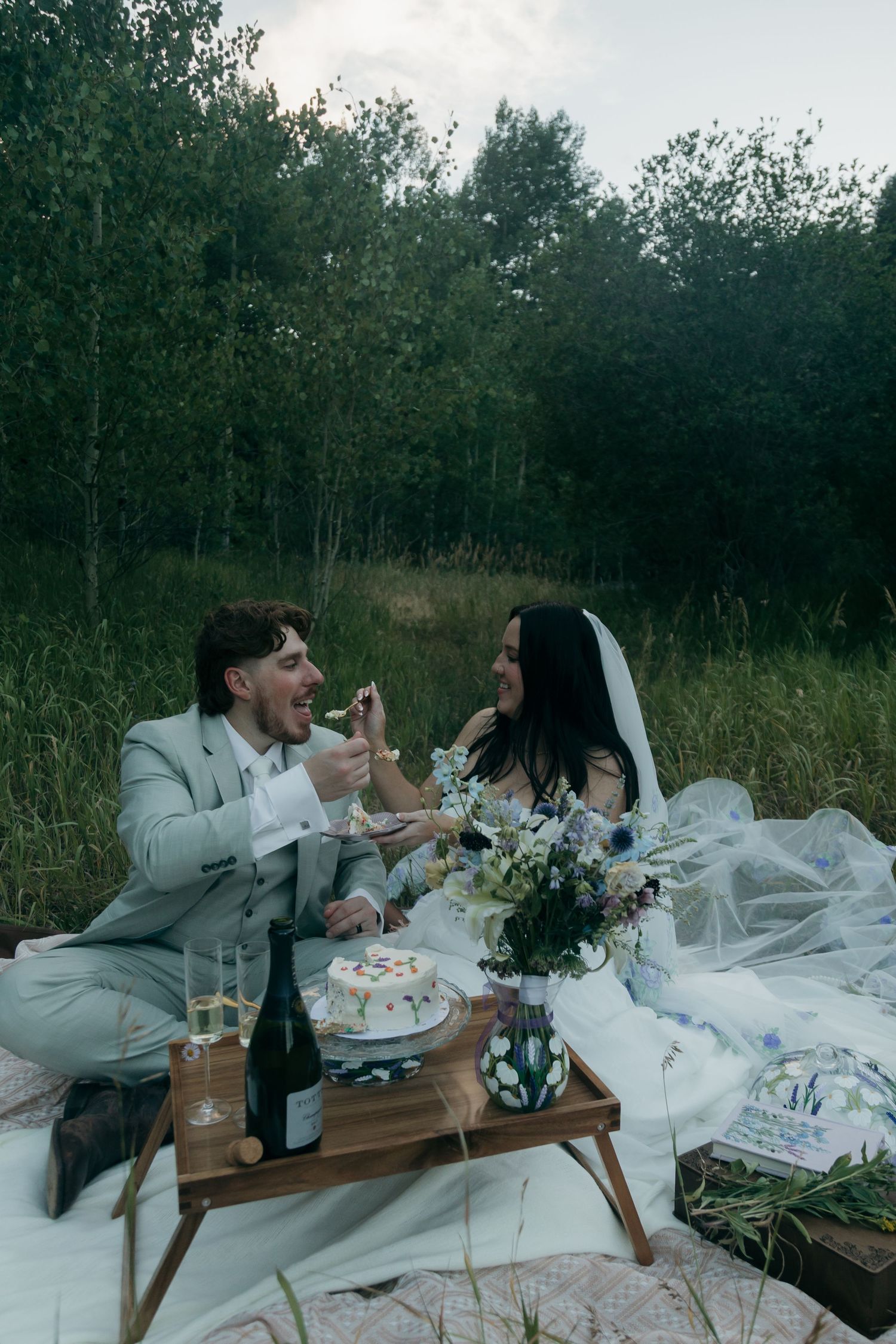 Newlyweds enjoy a romantic outdoor picnic with wedding cake and champagne on a white blanket in a forest meadow.