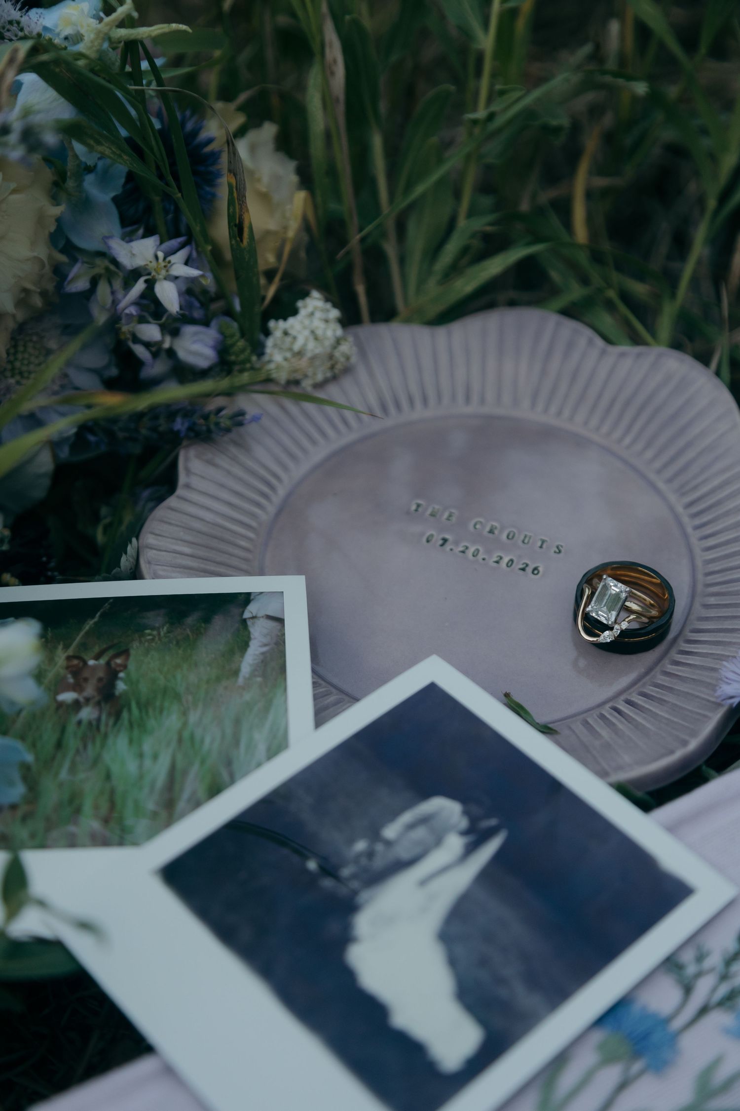 Wedding flat lay with rings on clay dish, Polaroids, and blue floral bouquet.