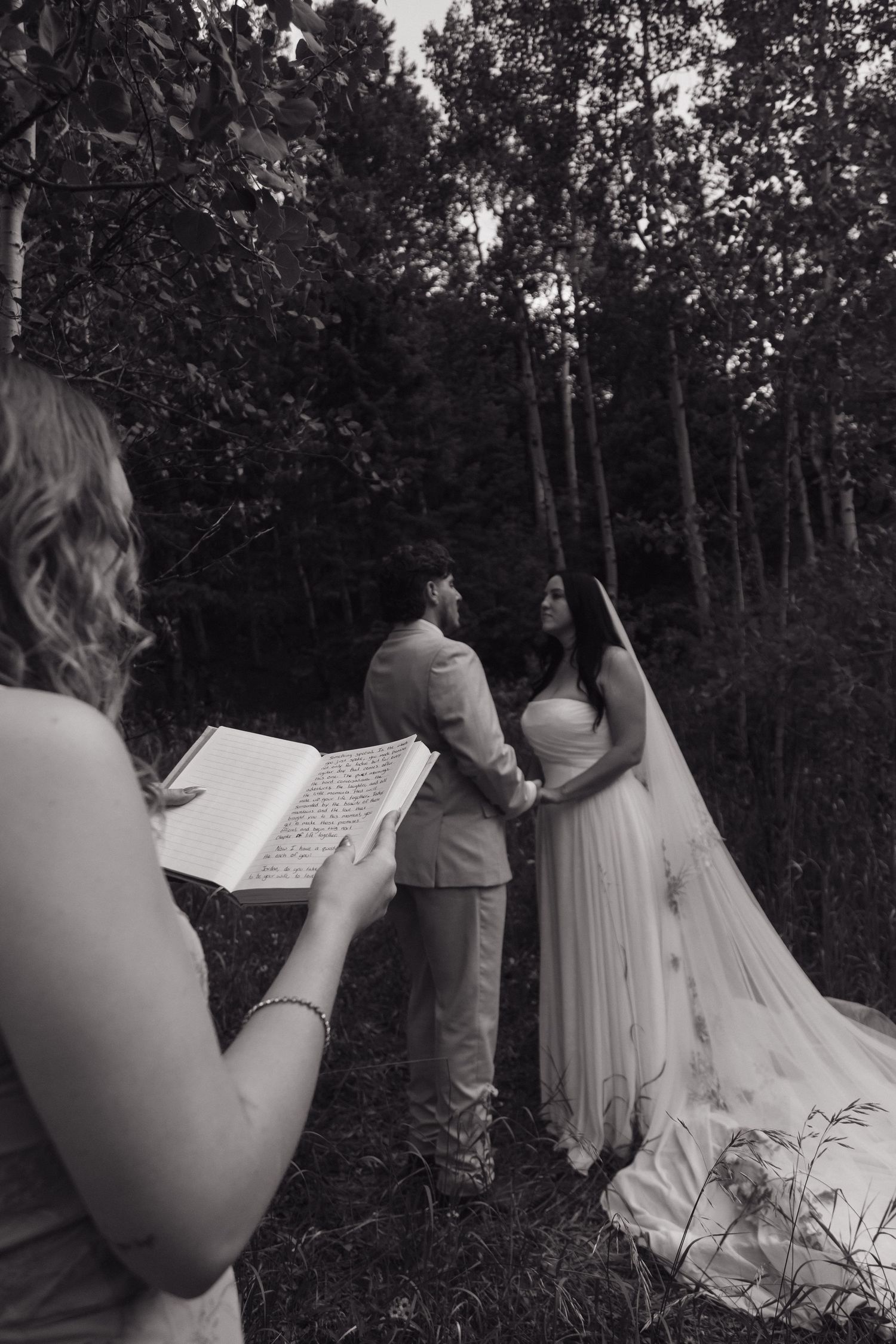 Black and white photo of an officiant reading from a book during an intimate forest wedding ceremony.