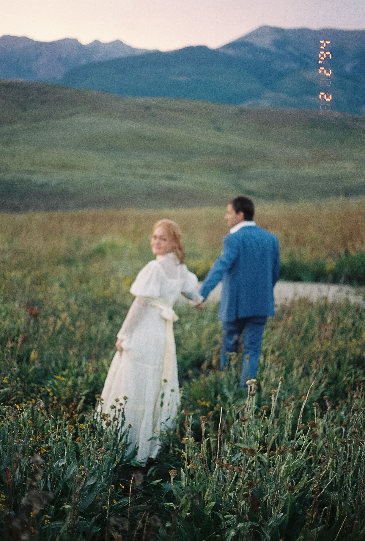 A bride in white dress holds grooms hand walking through a mountain meadow on film.