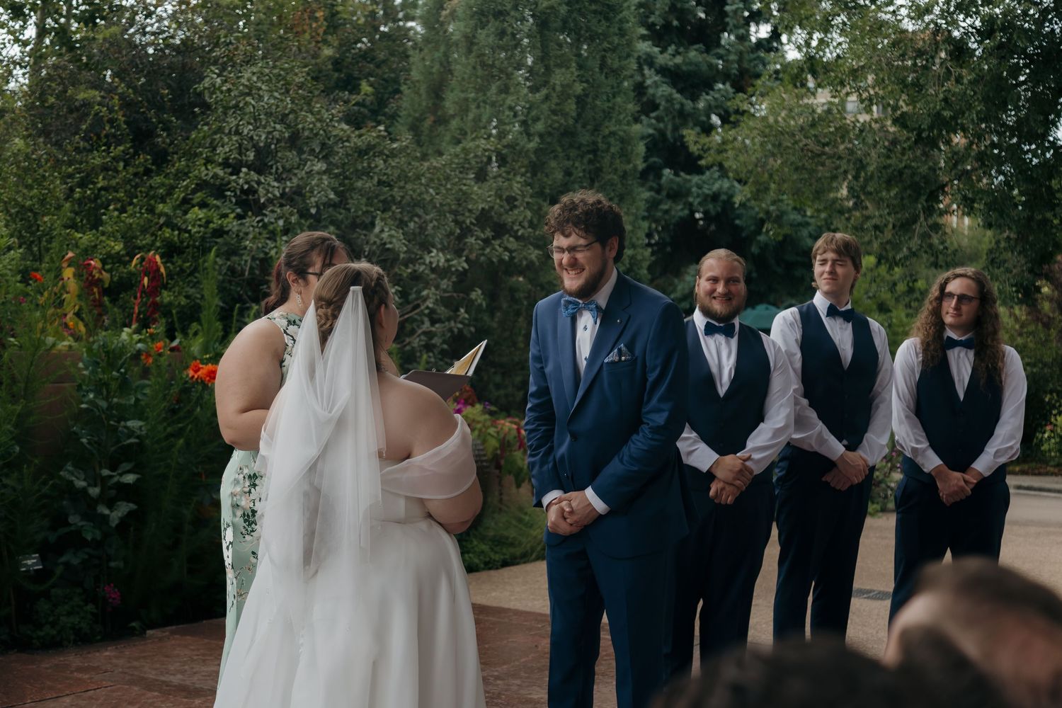 Bride reading vows to groom in blue suit at outdoor wedding ceremony with groomsmen standing behind.