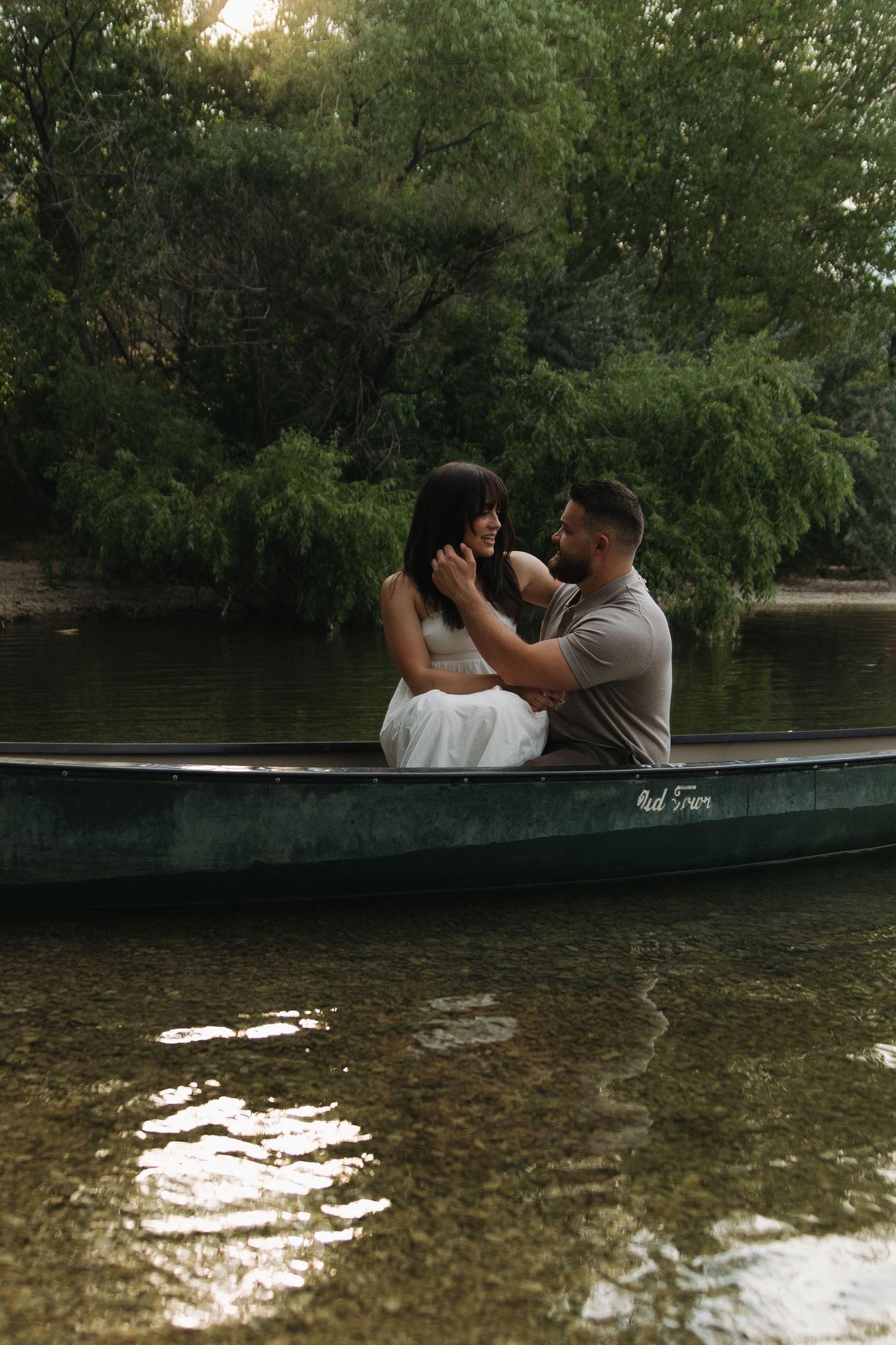 A couple shares a romantic moment in a canoe on a peaceful lake surrounded by green trees.