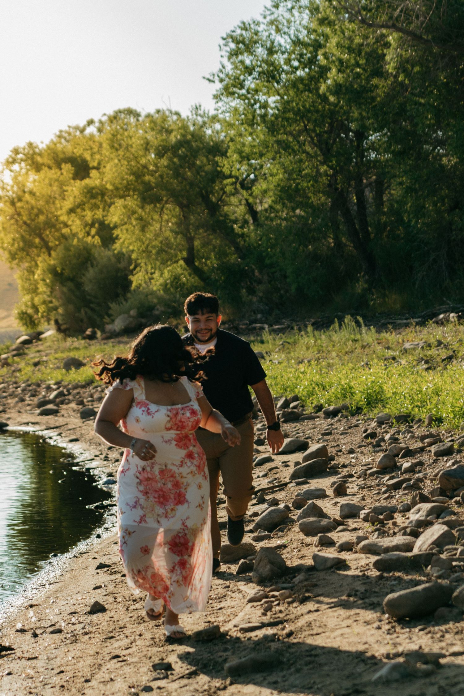 Couple in casual attire enjoying romantic moments along a rocky riverside during golden hour.
