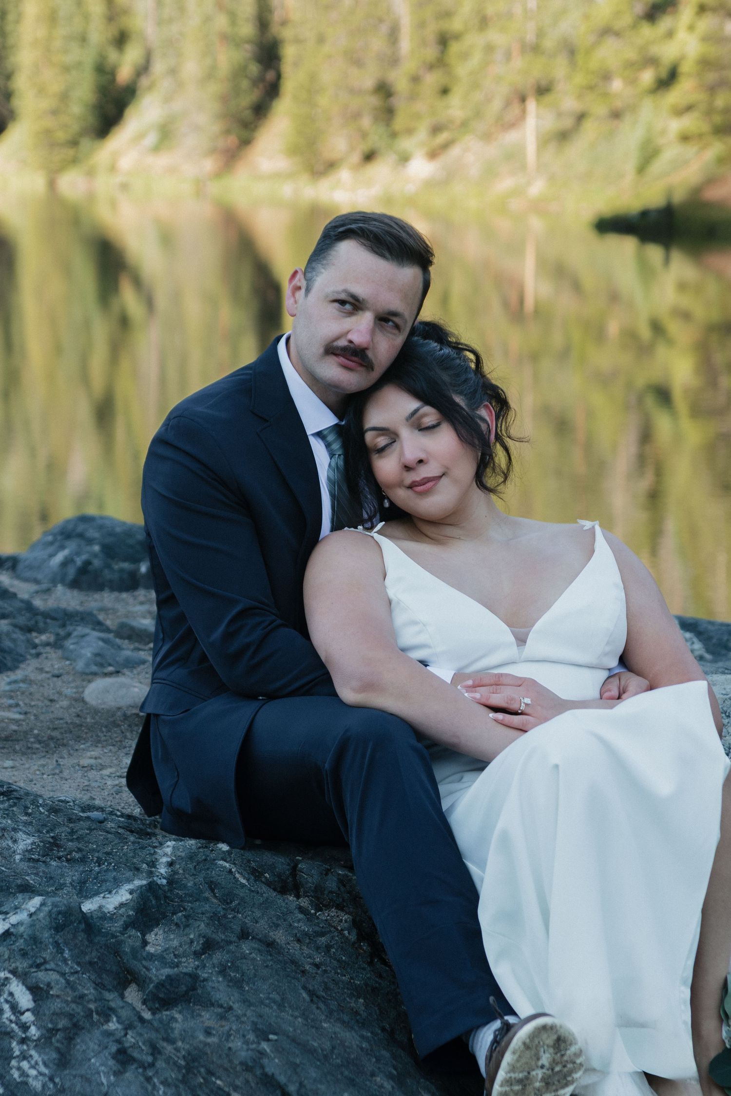 Couple rests on rocks by lakeside during mountain wedding photoshoot.