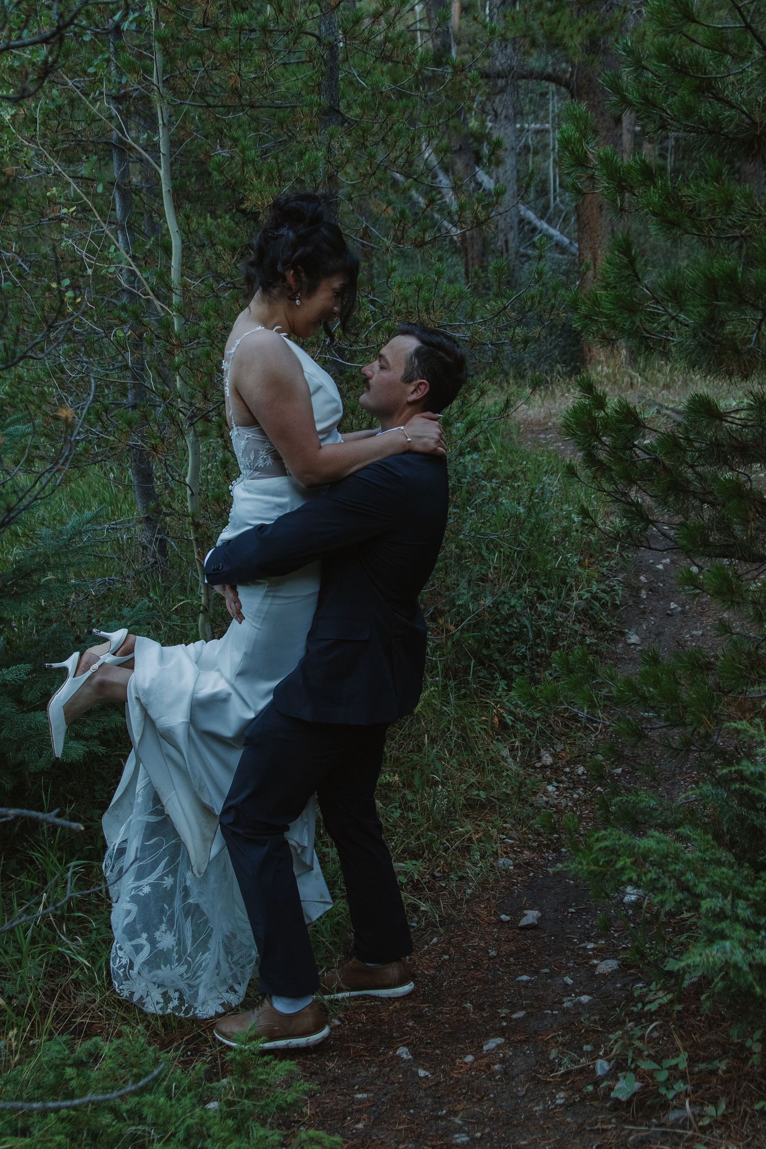 A couple in formal attire dances intimately on a wooded forest path during what appears to be a wedding celebration.