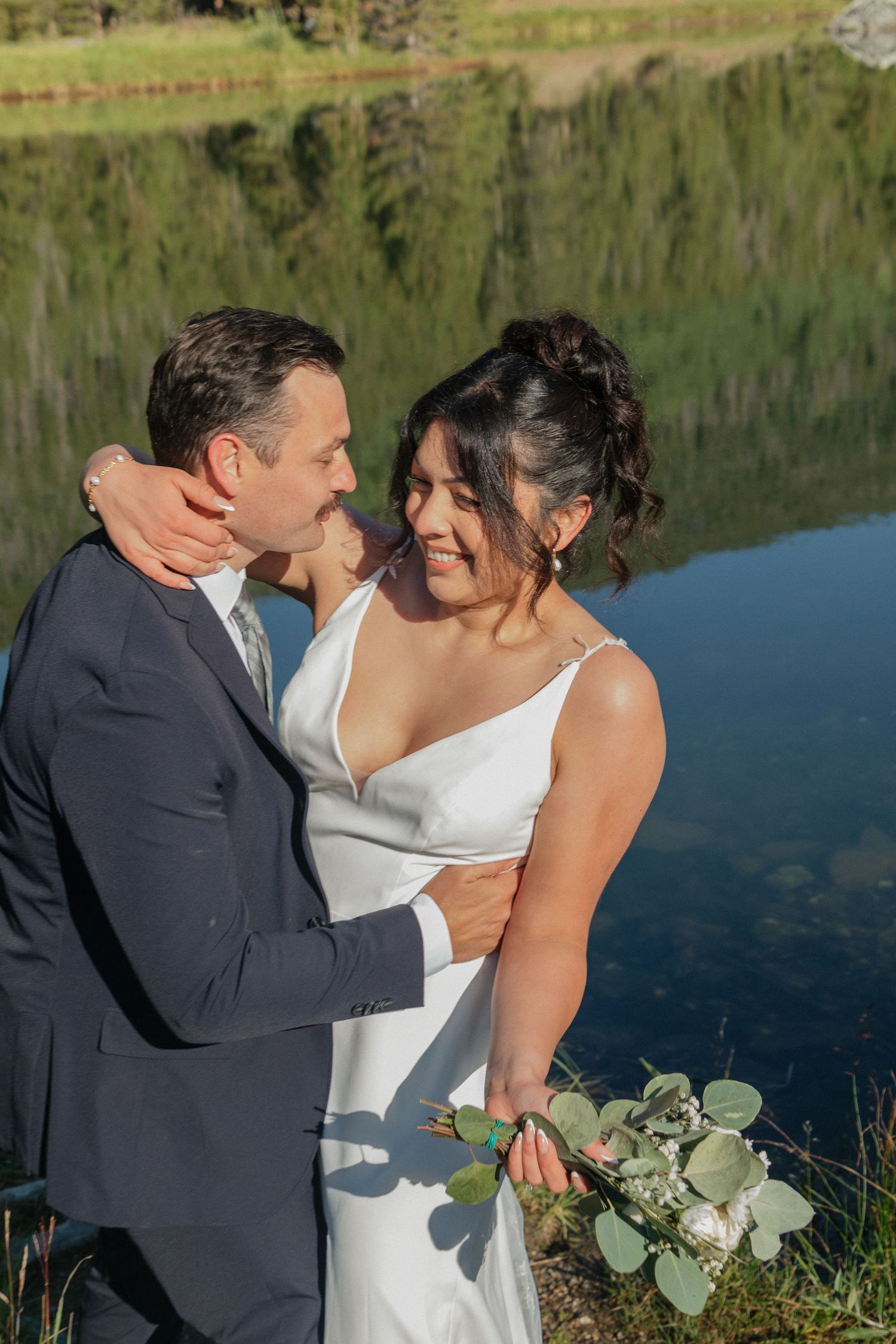 Sunny lakeside wedding portrait with reflective water and forest backdrop.
