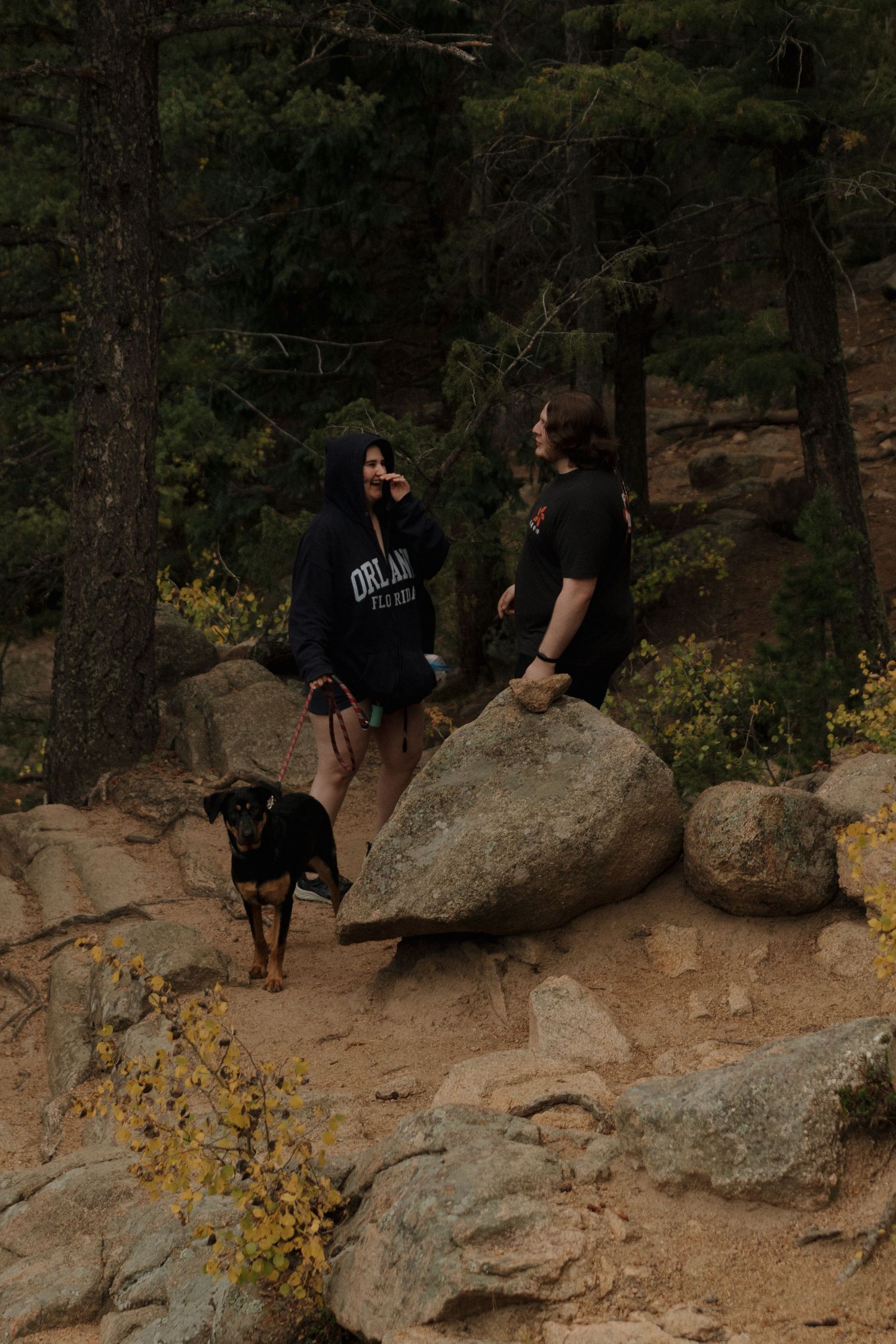 A couple stands on rocky terrain with their black dog while hiking through a forested area.