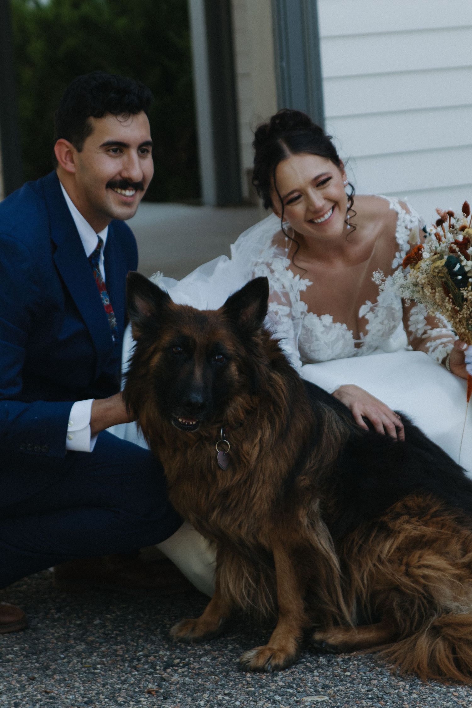 Wedding couple poses with their German Shepherd dog outside on a porch.