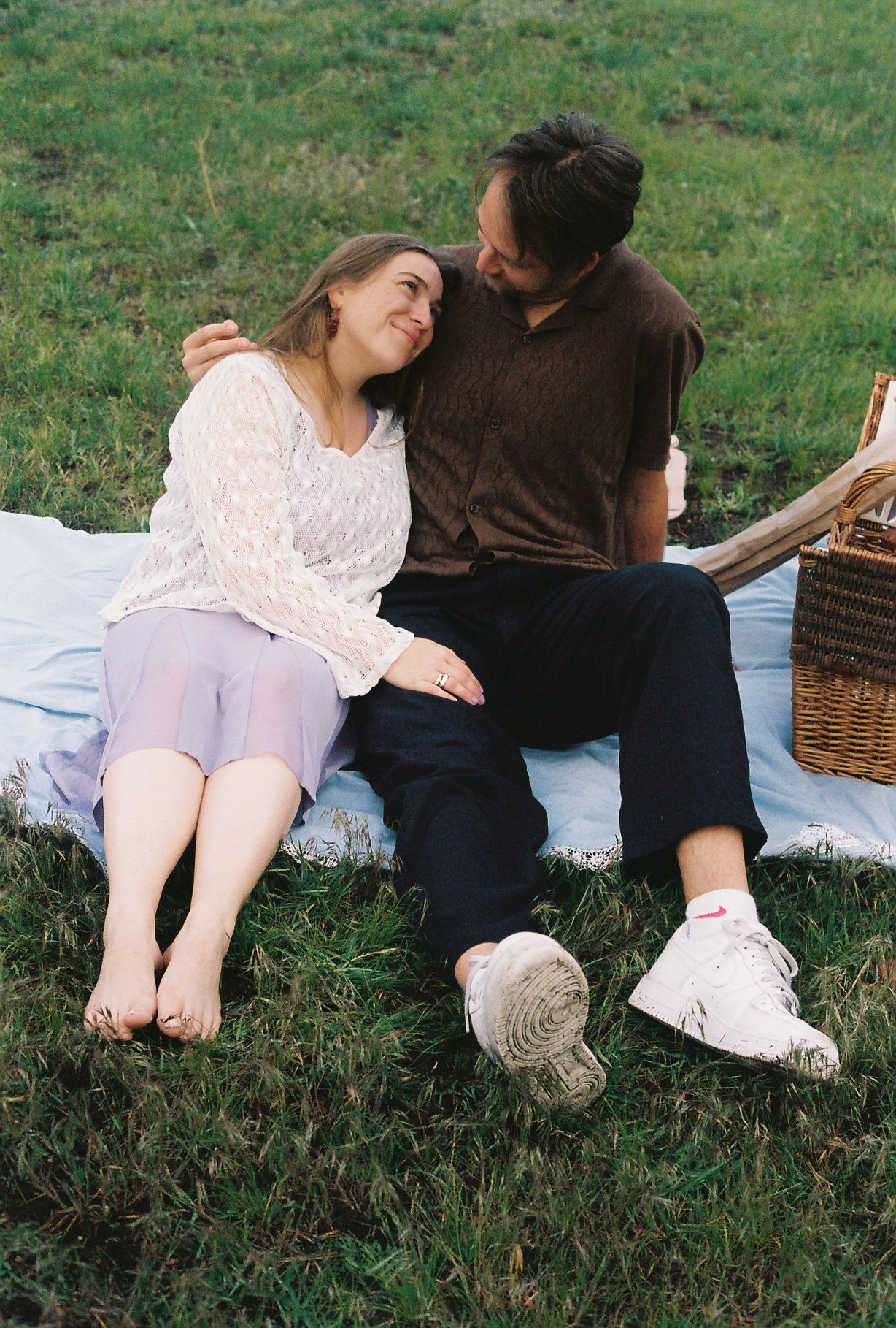 A happy couple sits together on a blanket in a grassy field during a picnic.