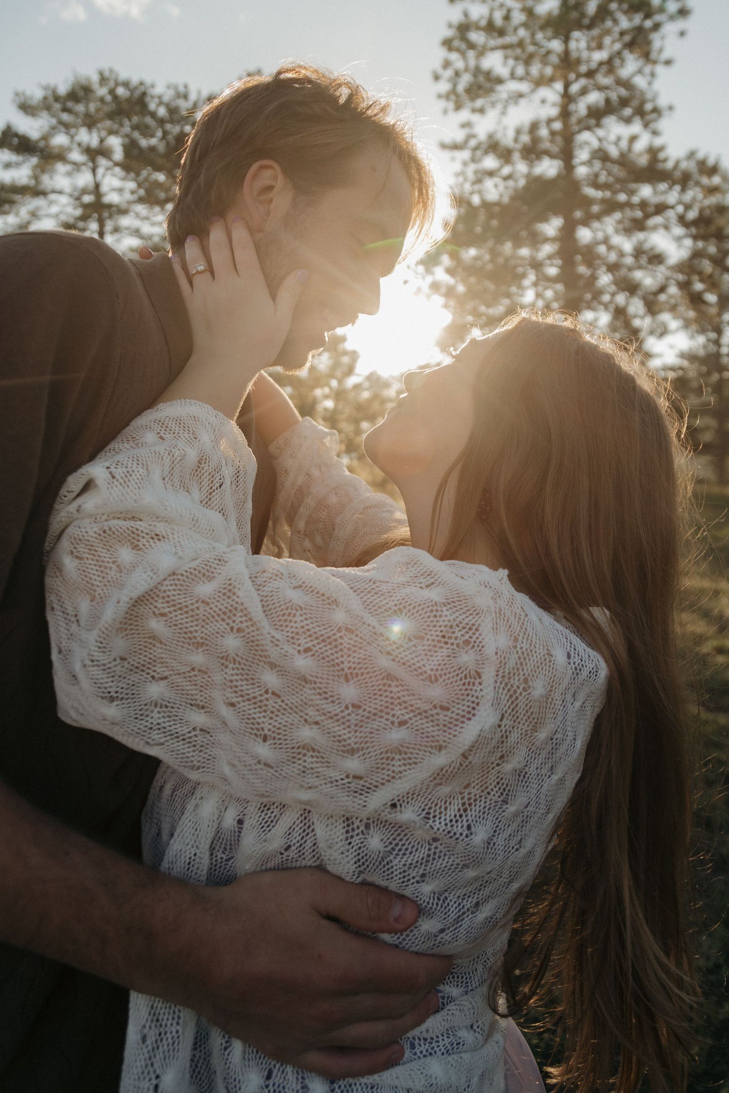 Couple sharing a golden sunset kiss outdoors, woman in white lace dress holding mans face tenderly.