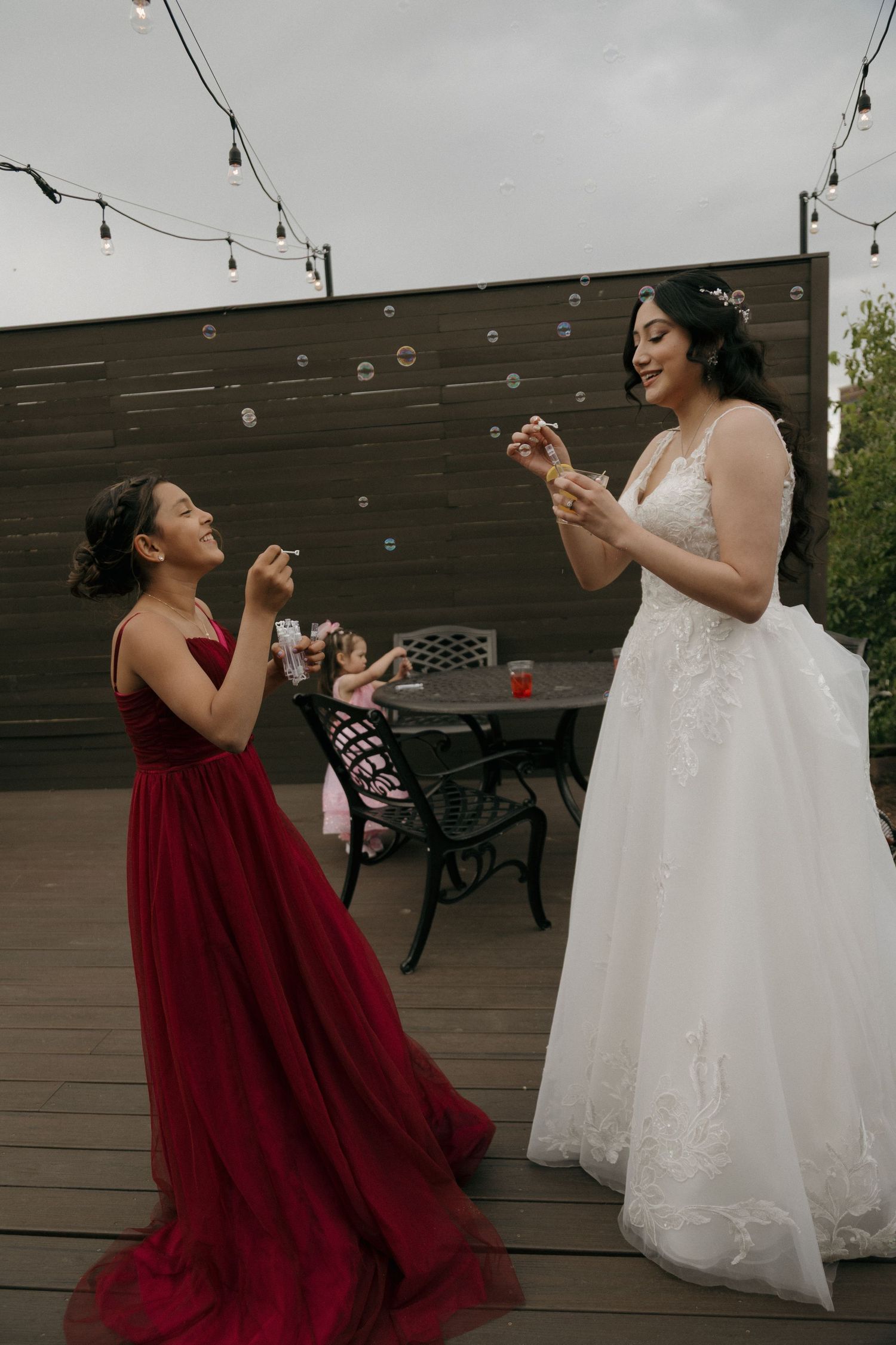Bride in white lace gown blowing bubbles with young girl in red dress at outdoor wedding.