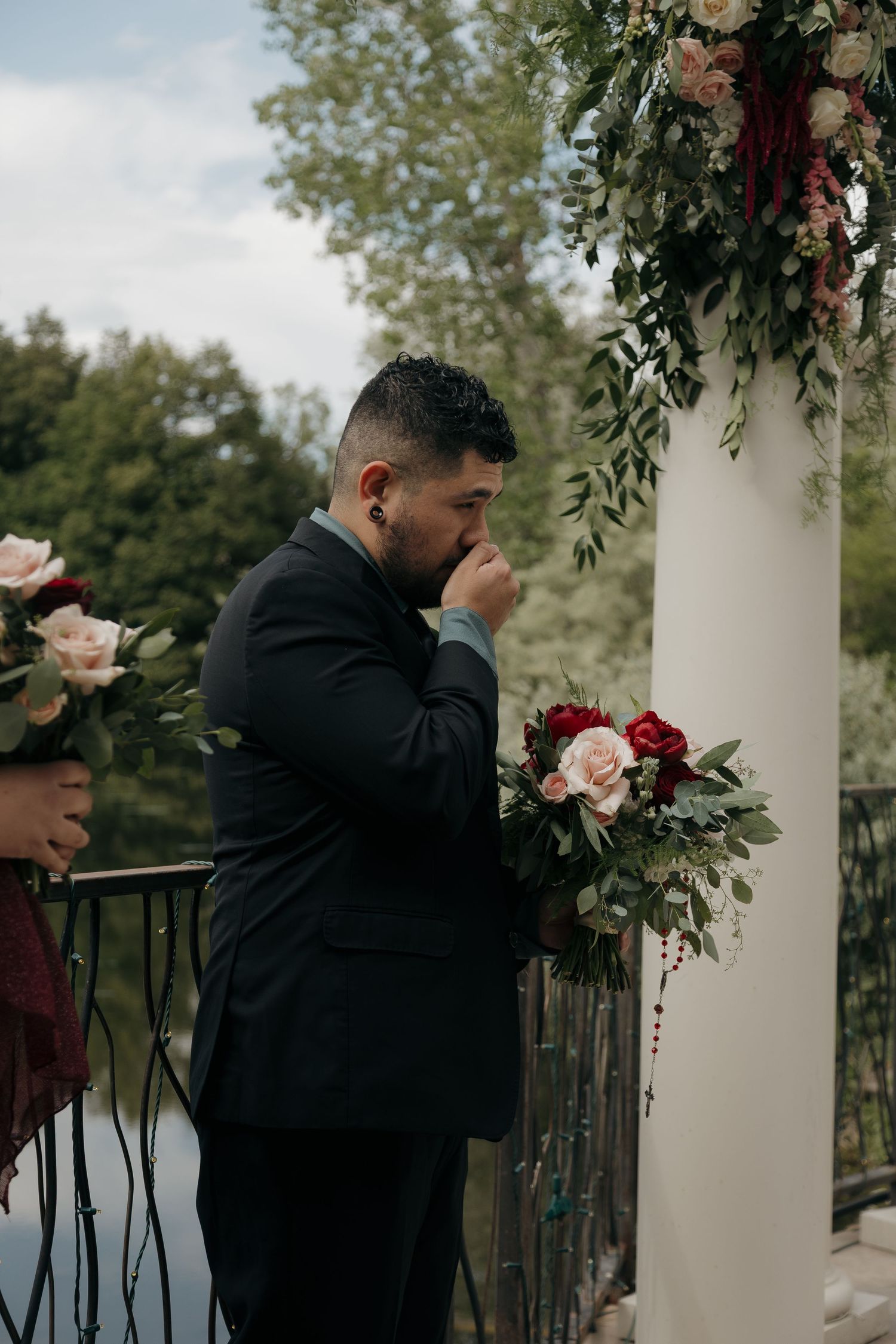 Emotional groom in black suit kisses bride's bouquet of red and blush roses during outdoor wedding ceremony.