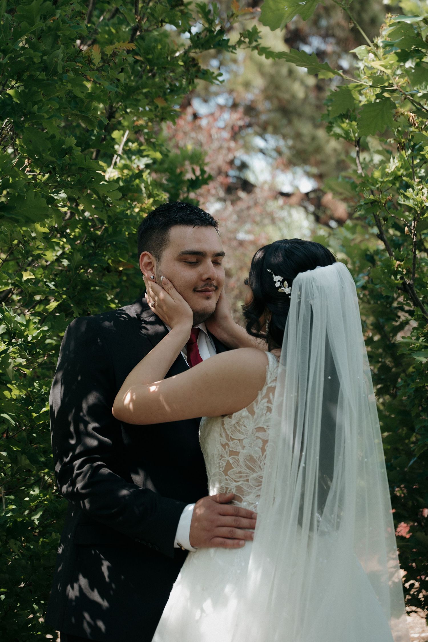 Groom tenderly holding bride's face for a kiss surrounded by green foliage on wedding day.