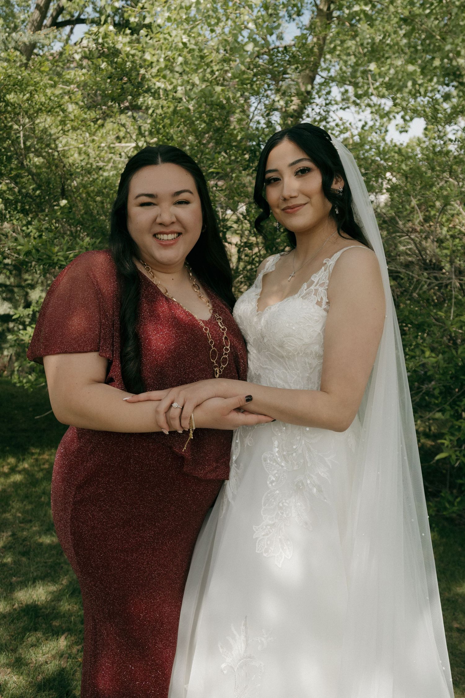 Bride in lace wedding gown with veil poses with bridesmaid in red dress outdoors.