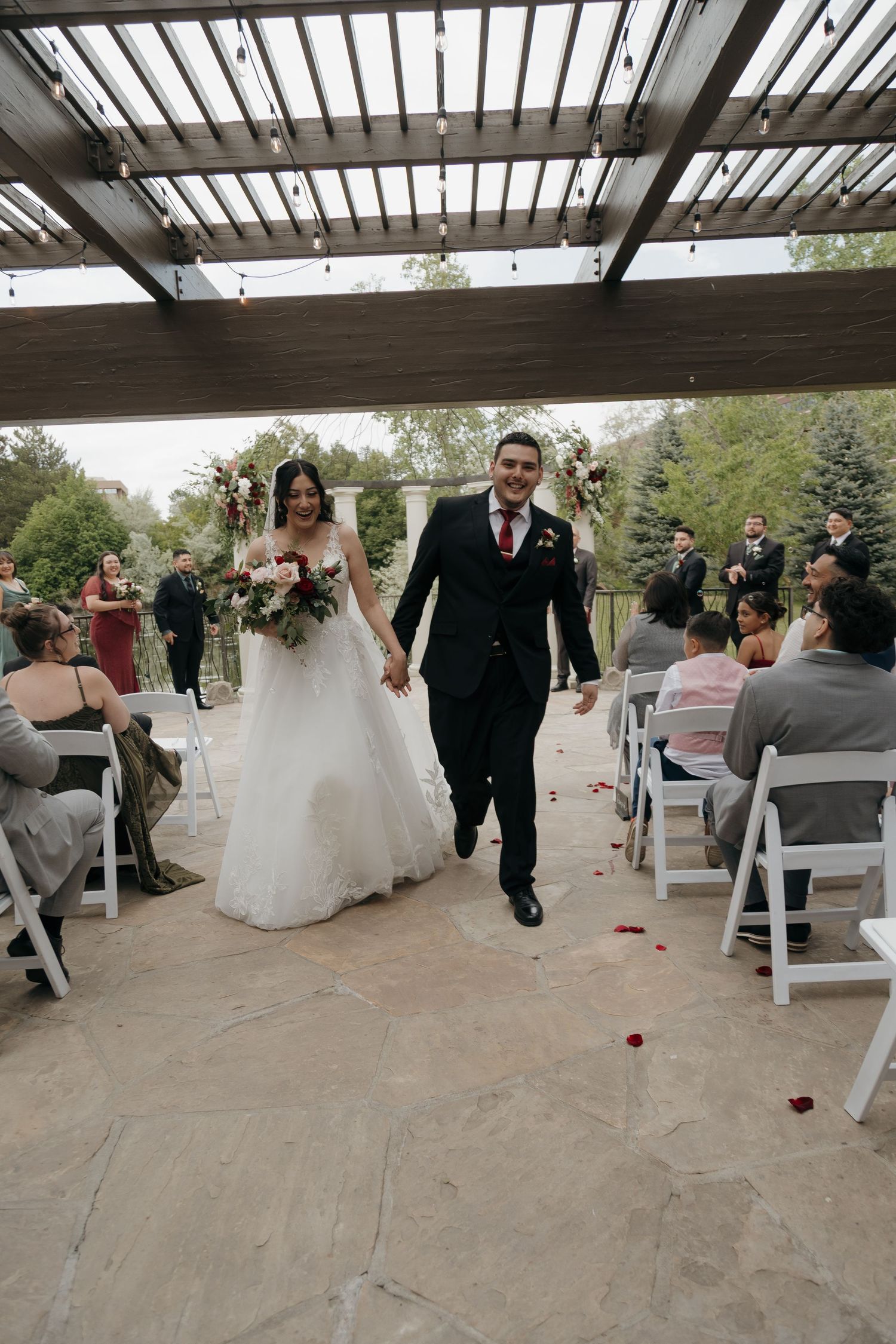 Joyful newlyweds walk back down the aisle after wedding ceremony under pergola with string lights and guests applauding.
